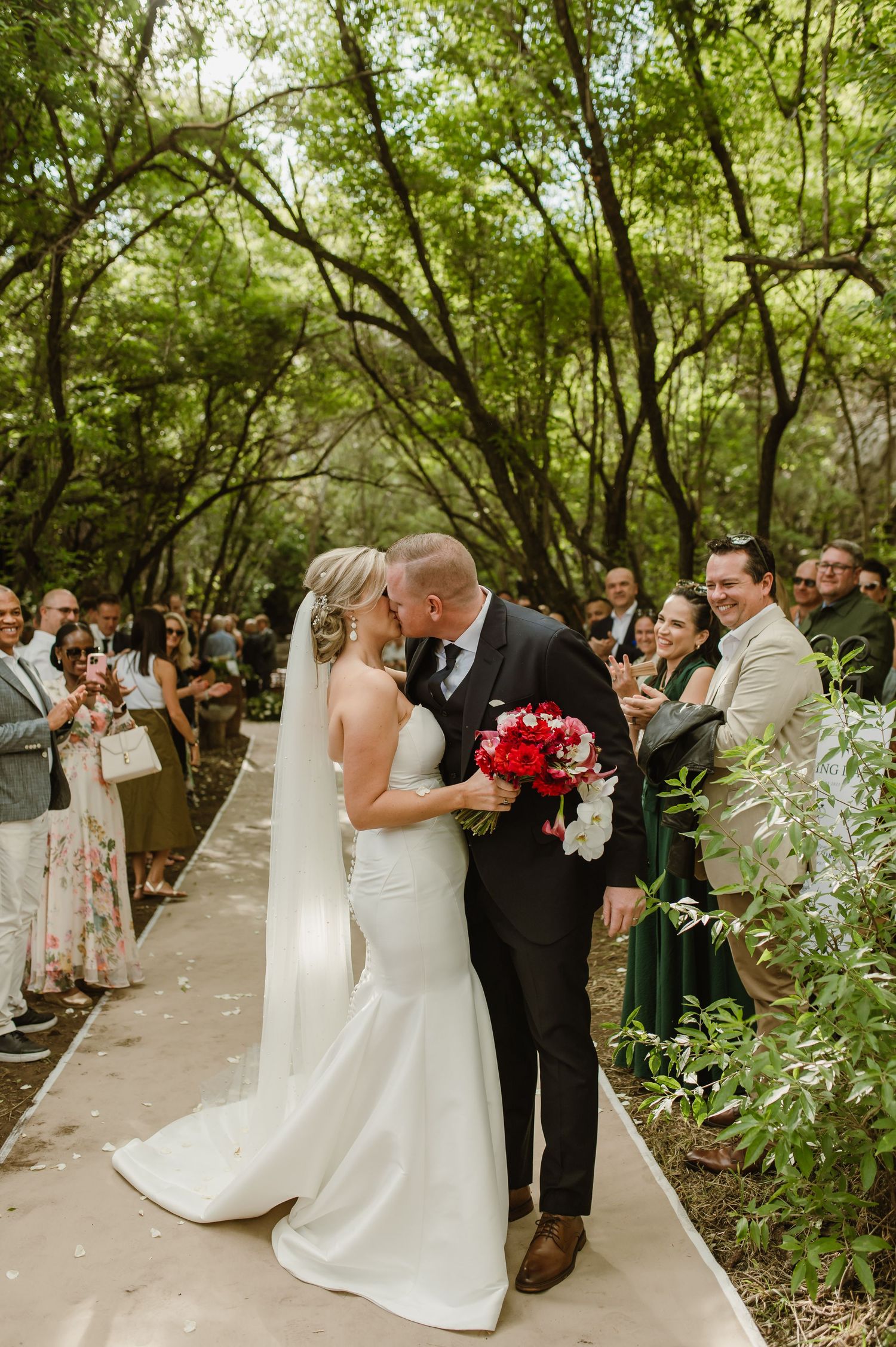 Wedding processional on a winding forest path with sunlight filtering through the trees.