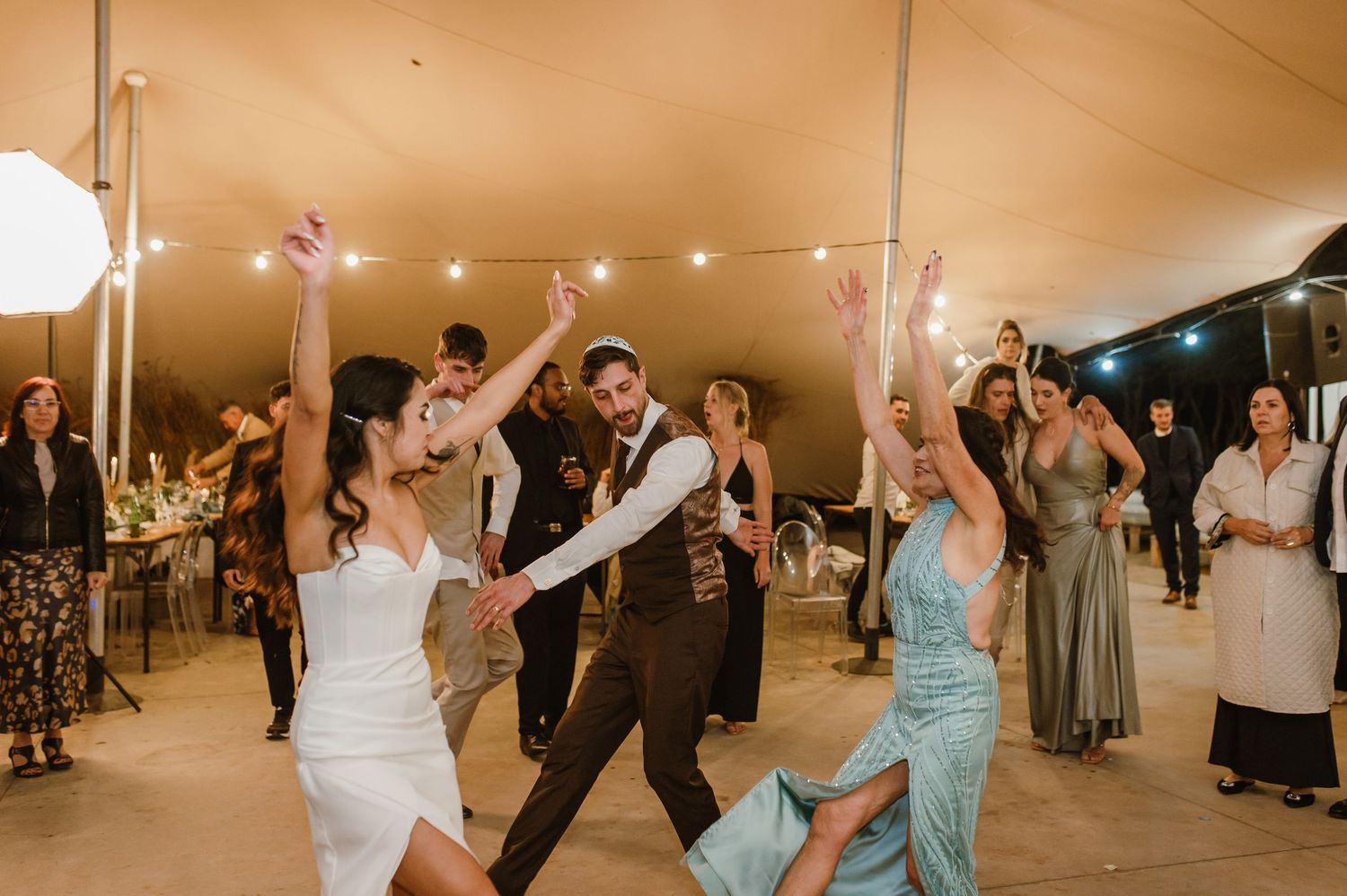 Wedding party members dance enthusiastically under a tent during an evening reception celebration.