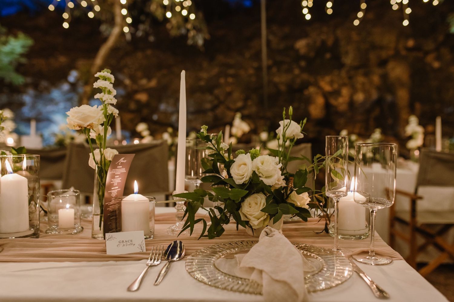 Elegant table settings with white linens, candles and greenery arrangements at a rustic outdoor venue.