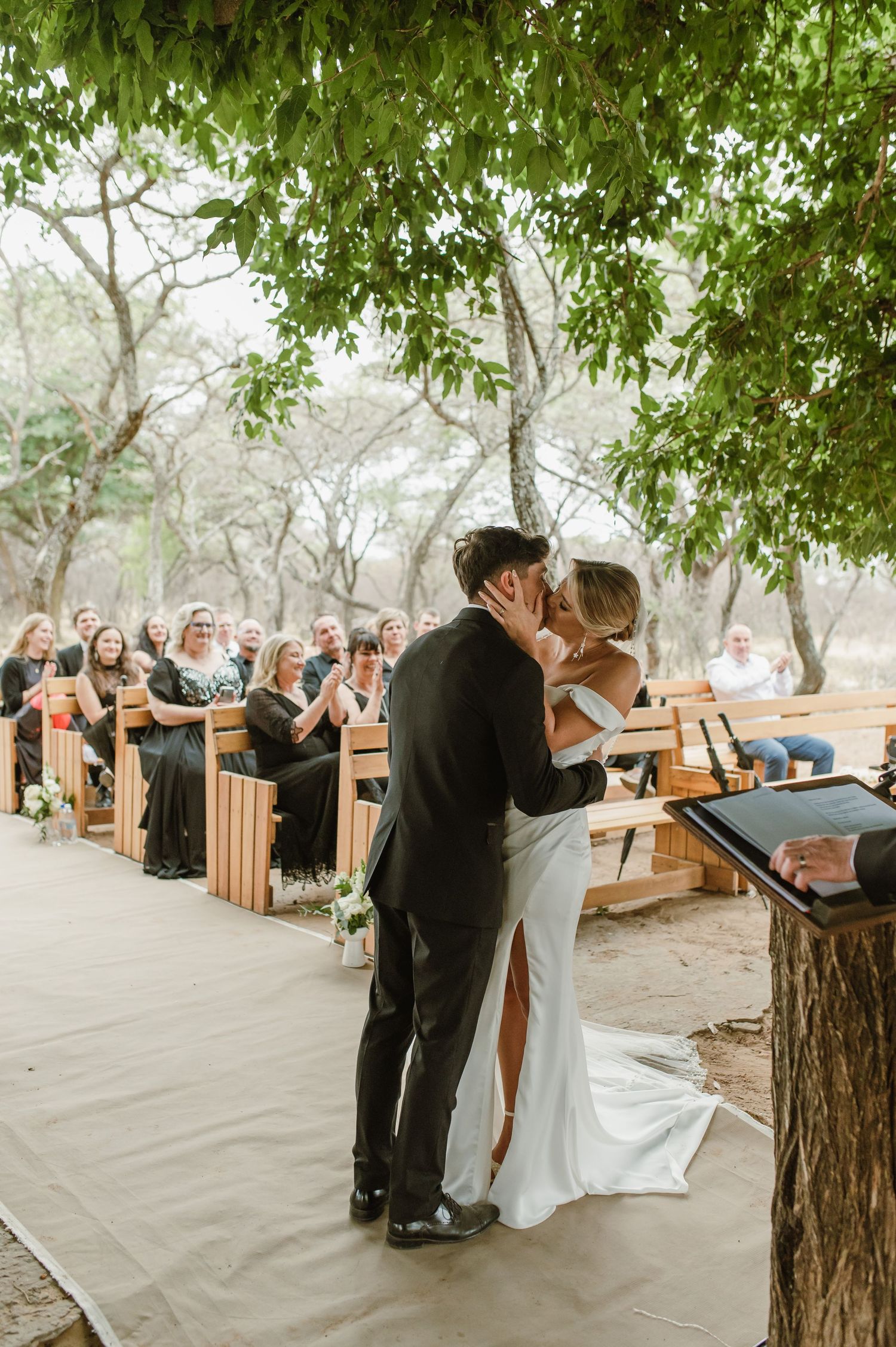 Newlyweds embrace during their rustic outdoor ceremony with wooden benches and leafy trees.