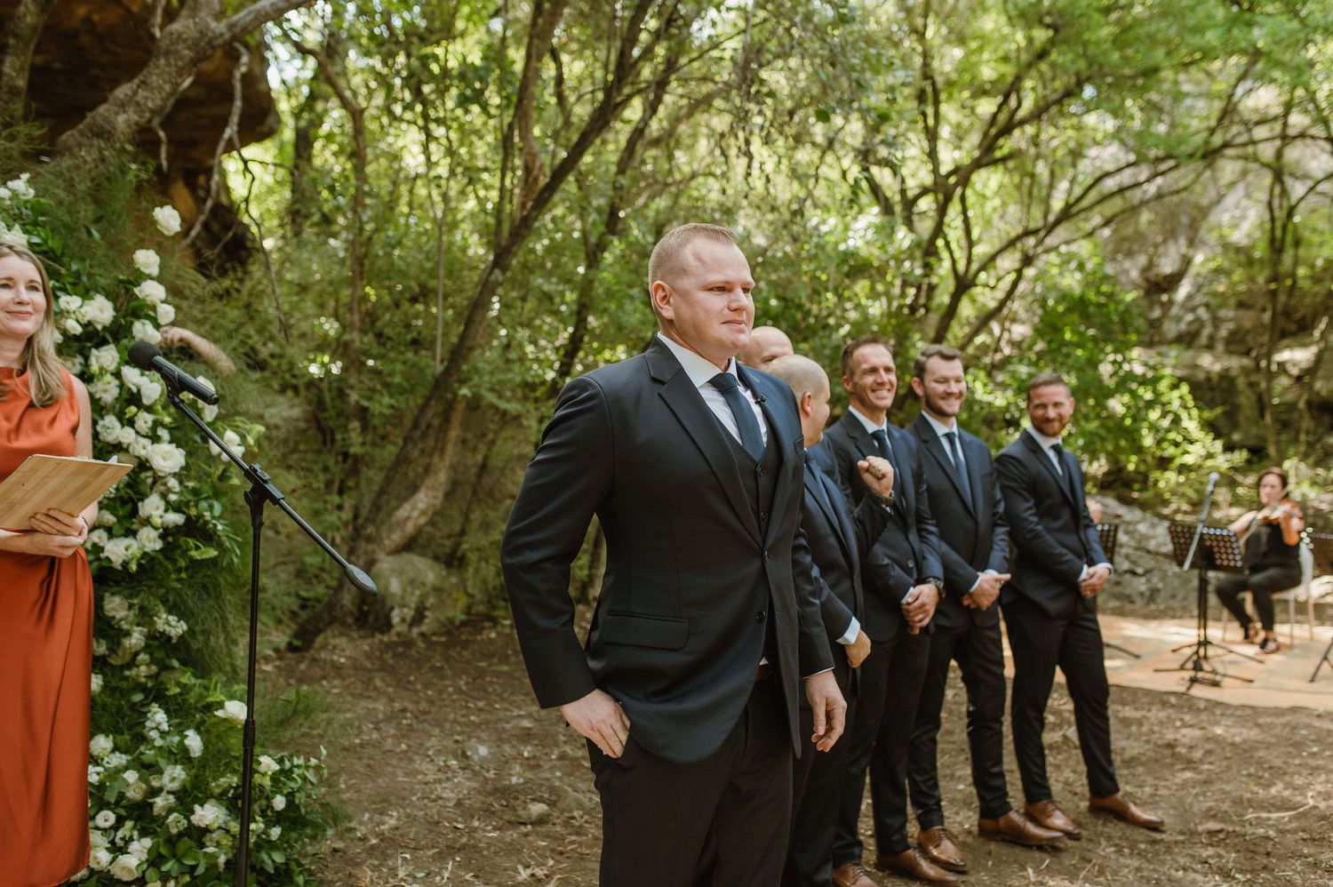 Groomsmen in dark suits stand in line during an outdoor forest ceremony.