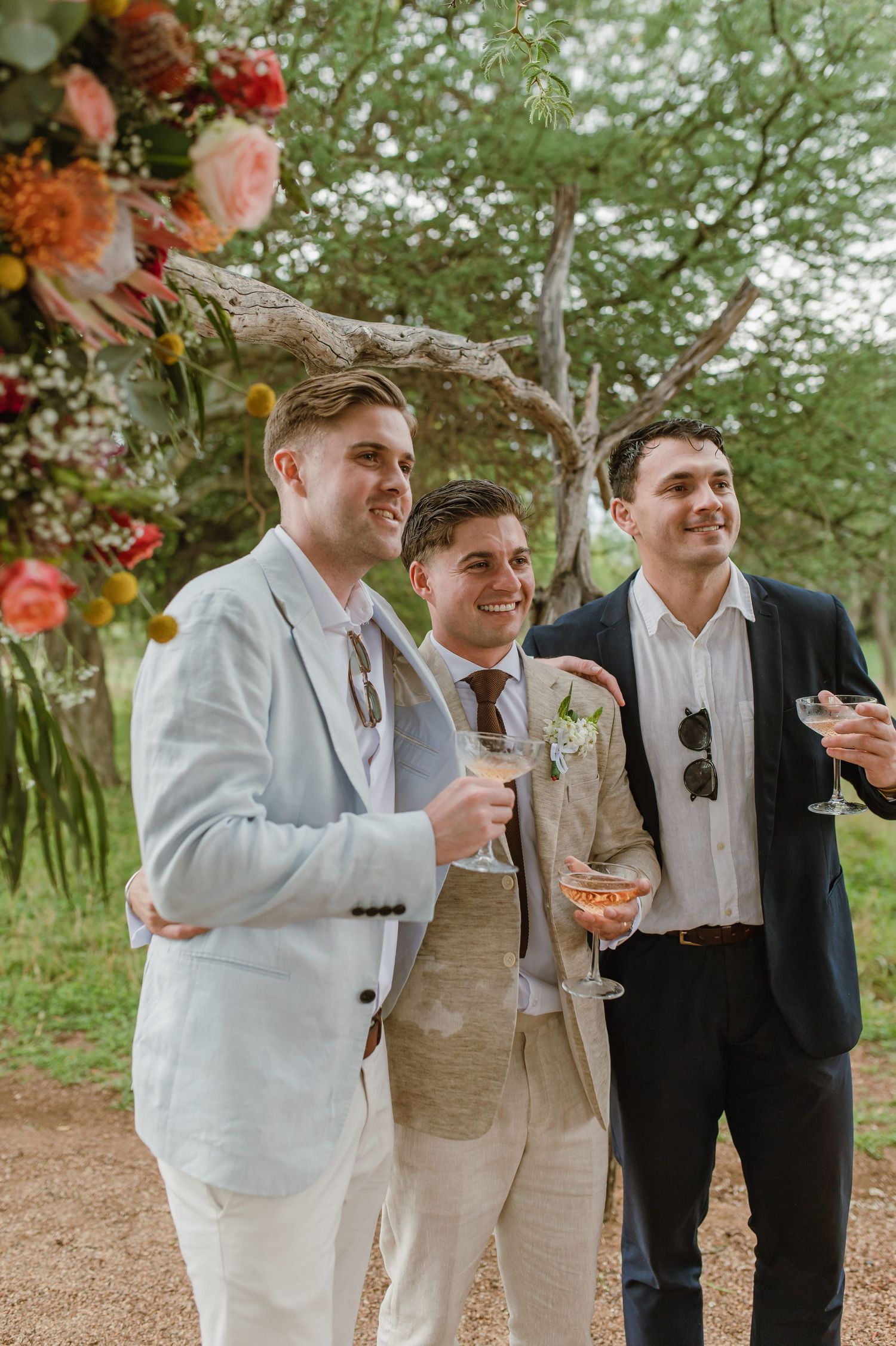 Groomsmen in light suits and dark ties toast during outdoor wedding celebration.