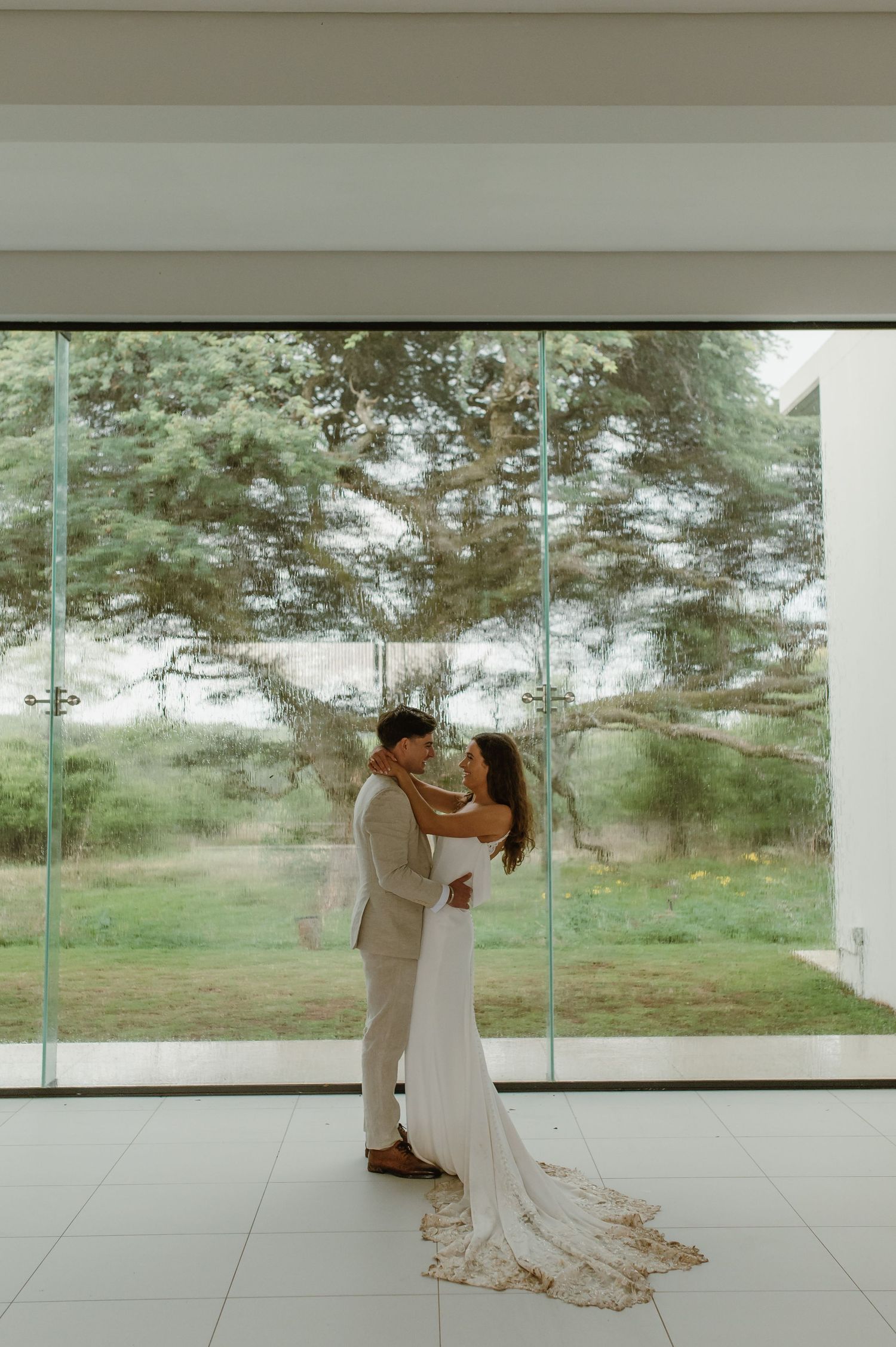 A romantic wedding portrait in a minimalist white room with floor-to-ceiling windows.