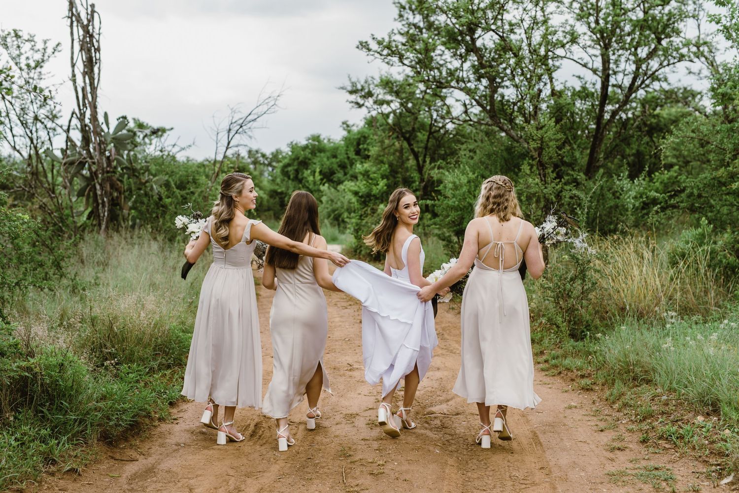 Bridal party in white and beige dresses walk together on outdoor path surrounded by greenery.