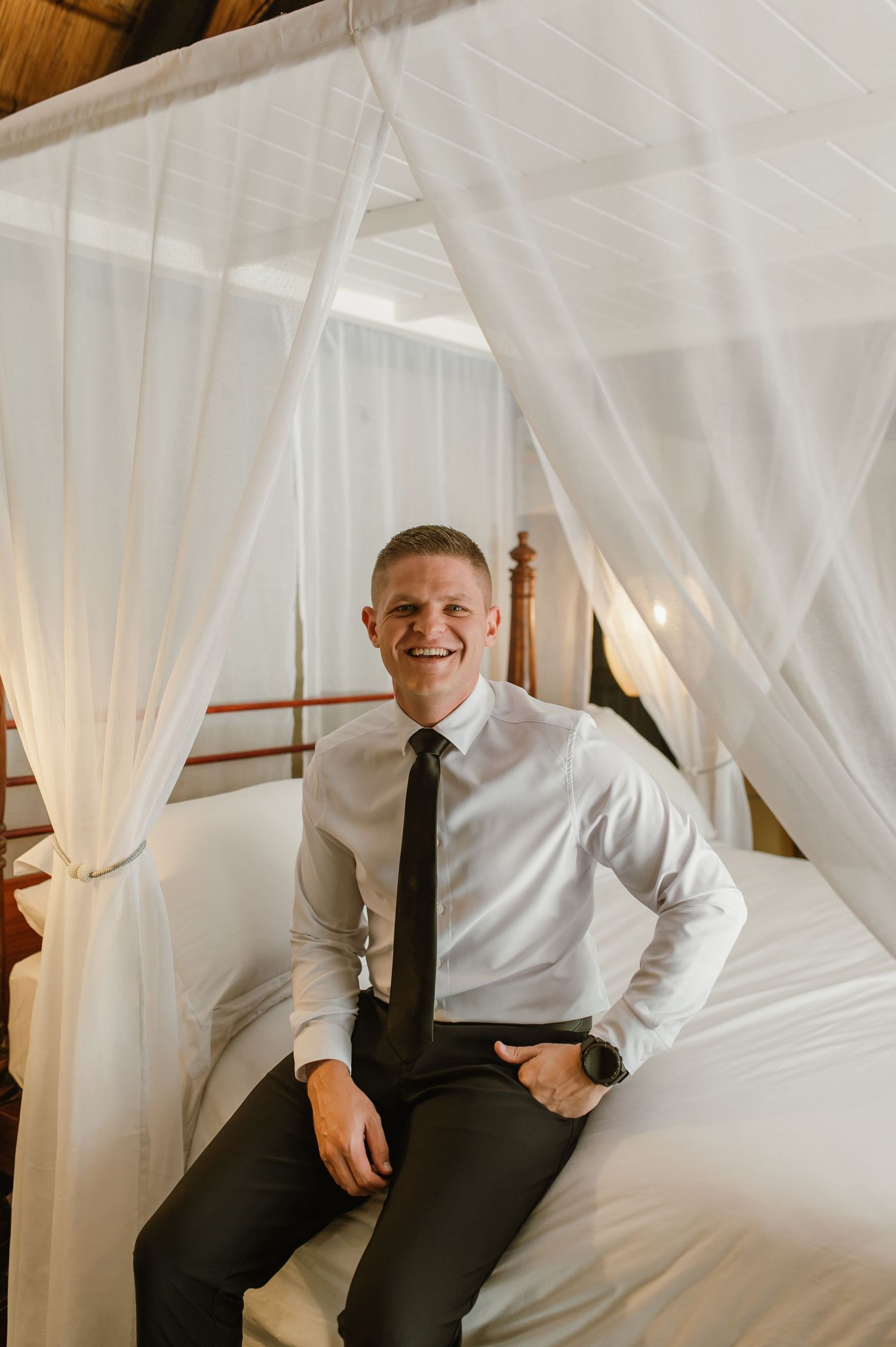 Smiling groom in white shirt and black tie seated on a four-poster canopy bed in safari lodge.