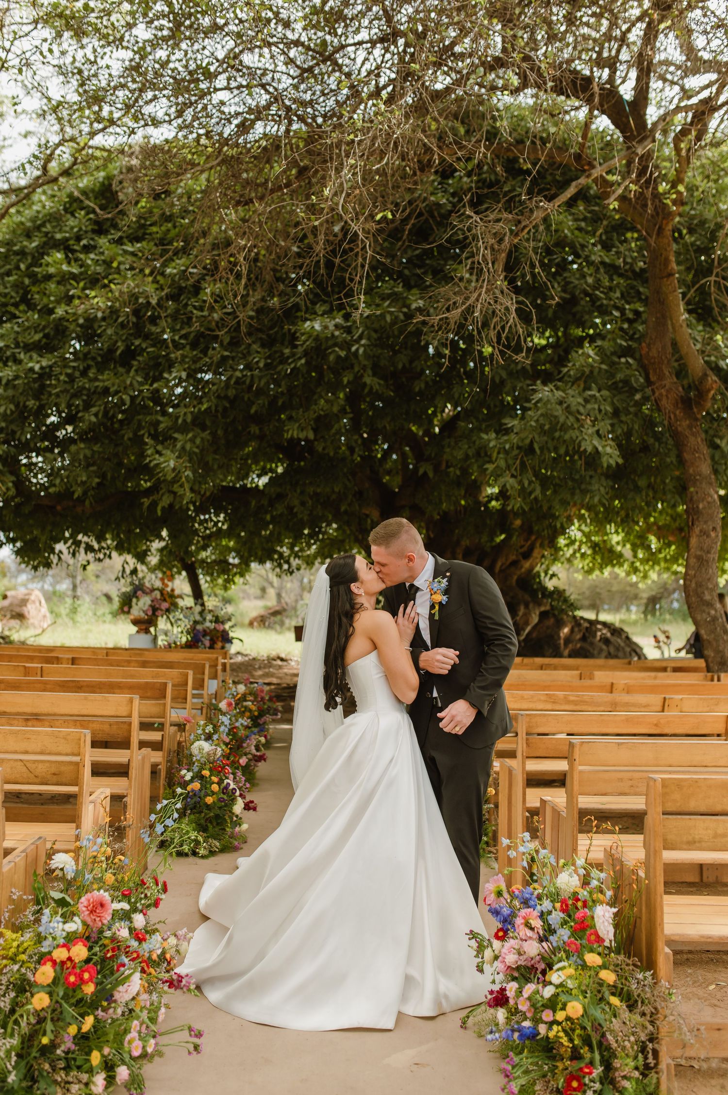 Bride and groom sharing first kiss at outdoor ceremony with colorful floral arrangements.