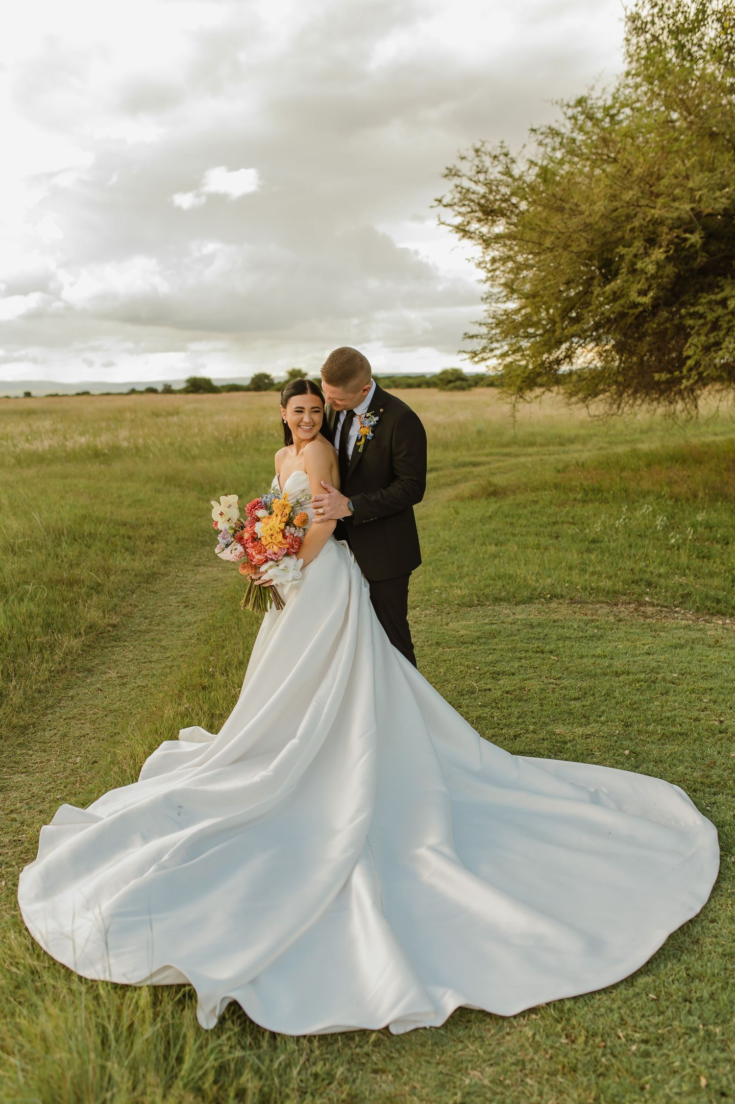Bride in stunning white gown with long train embraces groom in black suit in lush green field.