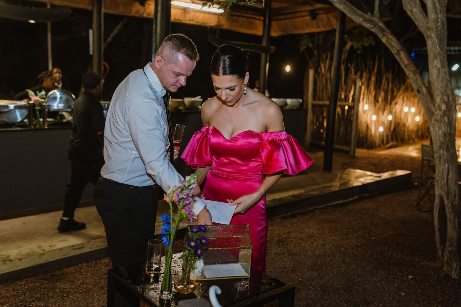 Woman in hot pink off-shoulder gown and man in blue shirt reviewing items at an evening reception.