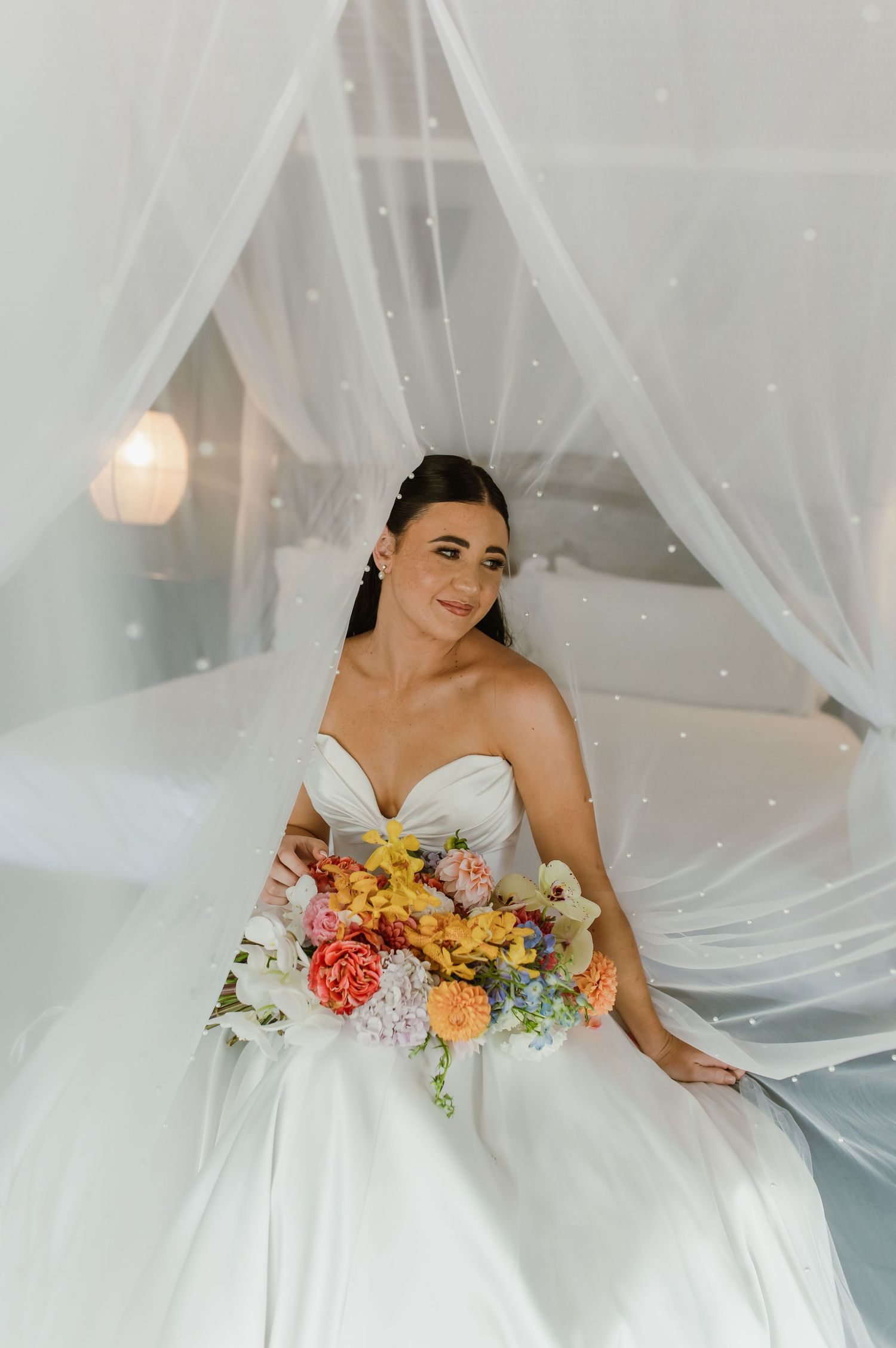 Bride in a strapless ballgown holding a vibrant colorful bouquet while seated under a white canopy bed.