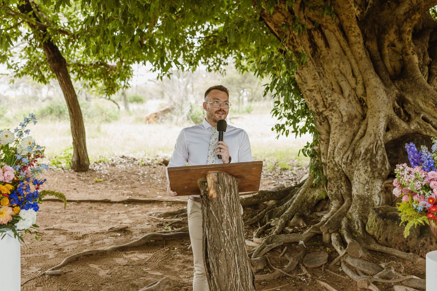 Smiling officiant speaking at an outdoor wedding ceremony beneath a large ancient tree with colorful flowers.