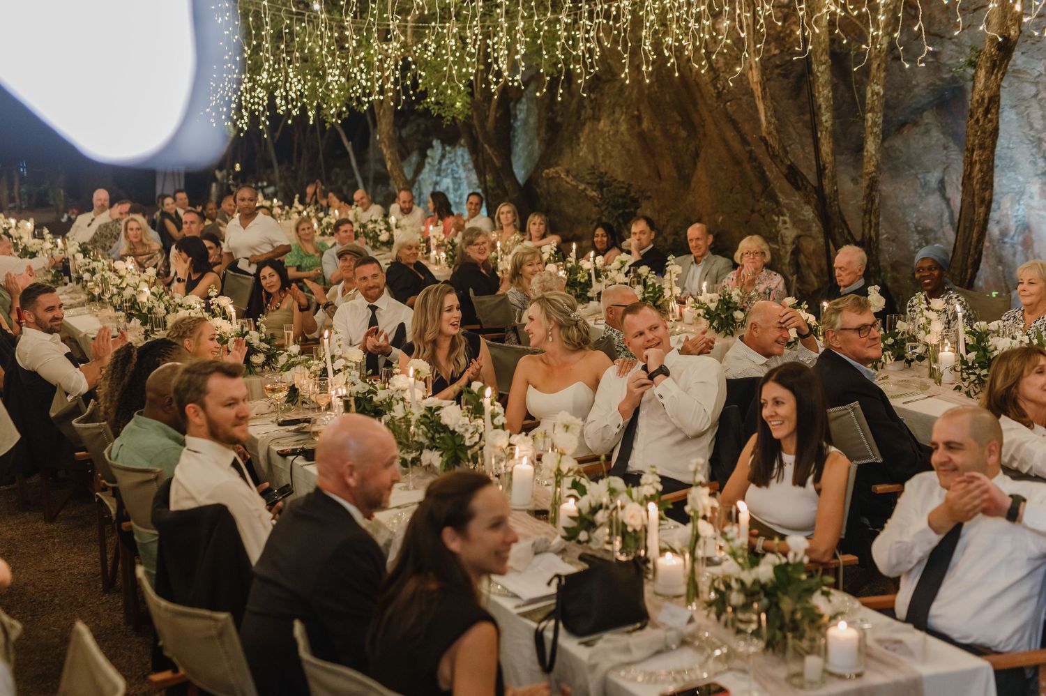 Wedding celebration with guests dining at elegantly decorated tables under warm lighting.