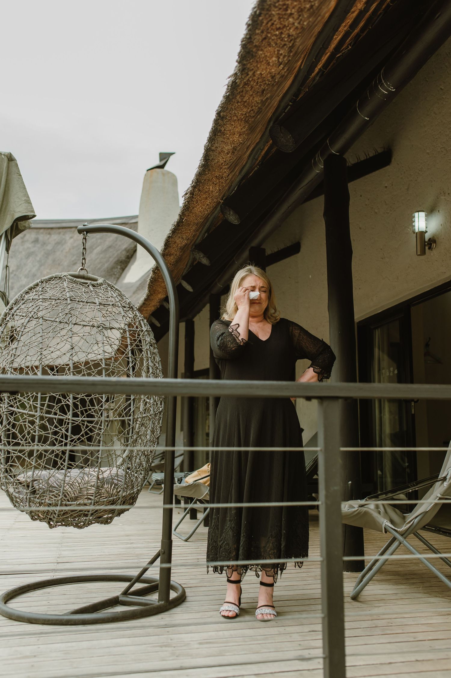 Woman in black dress stands on deck with hanging egg chair and thatched roof above.