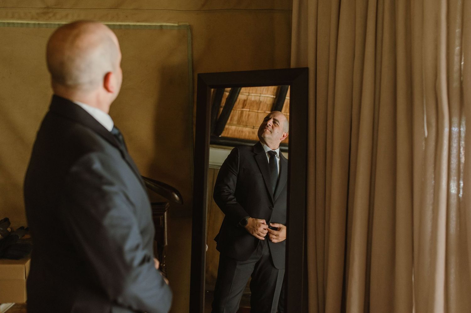 Men in formal suits getting dressed in a dimly lit room with curtains.