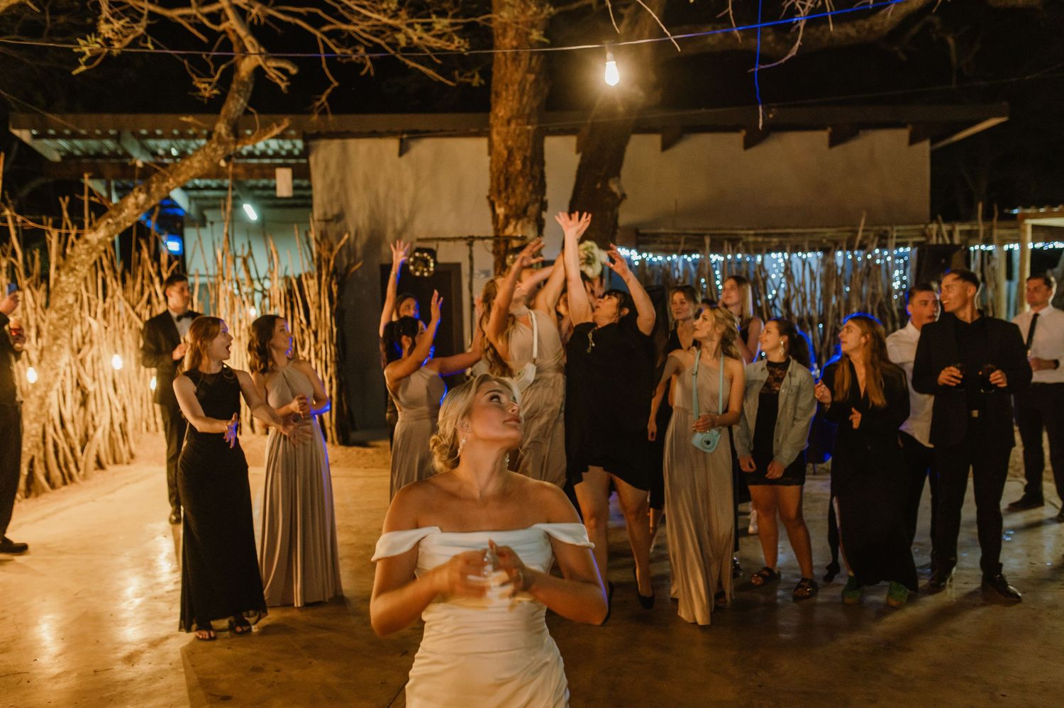 Wedding party and guests gathered in rustic venue with overhead lighting and natural elements.