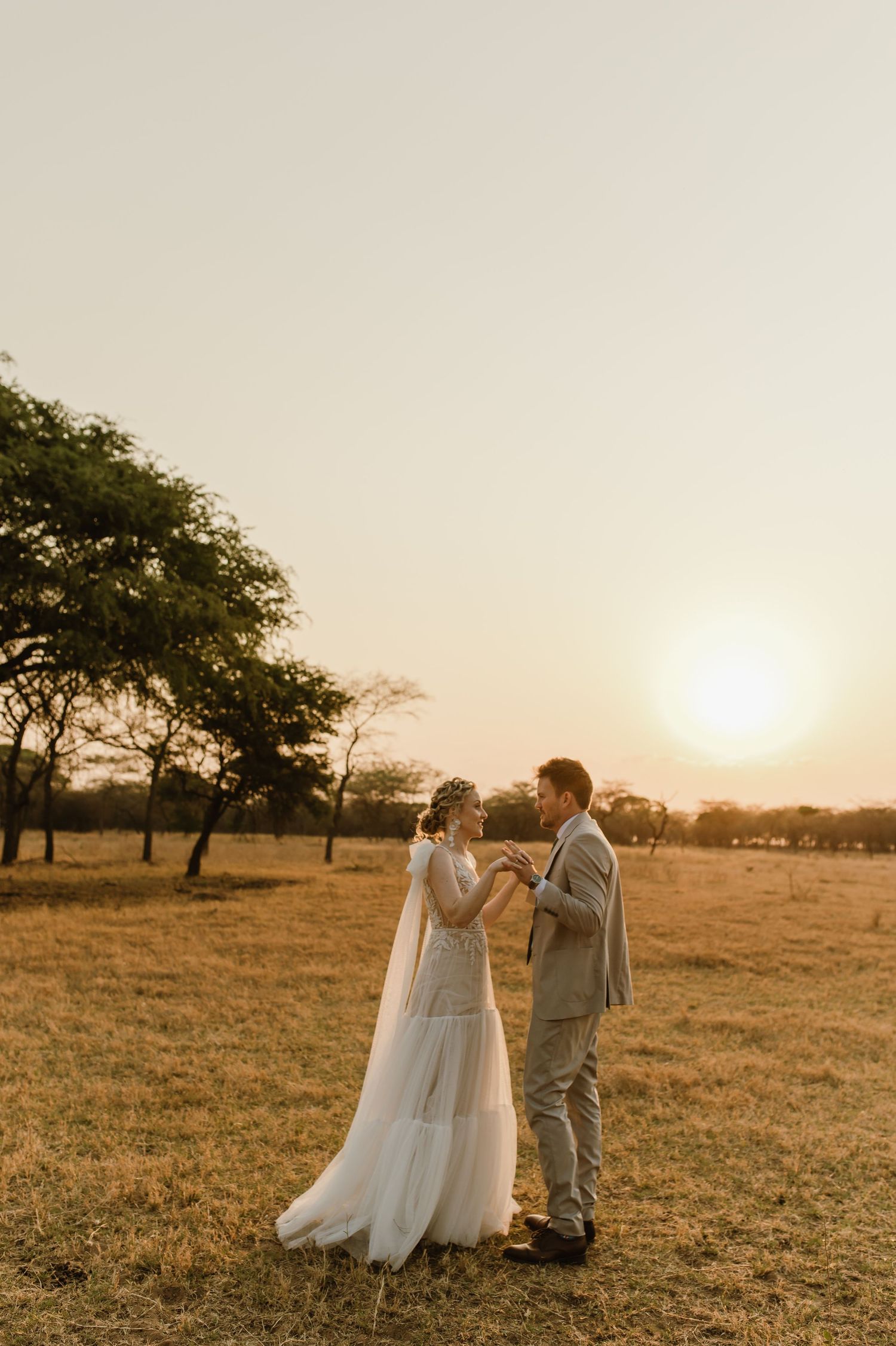 A couple in wedding attire stands together in a golden field as the sun sets behind them.