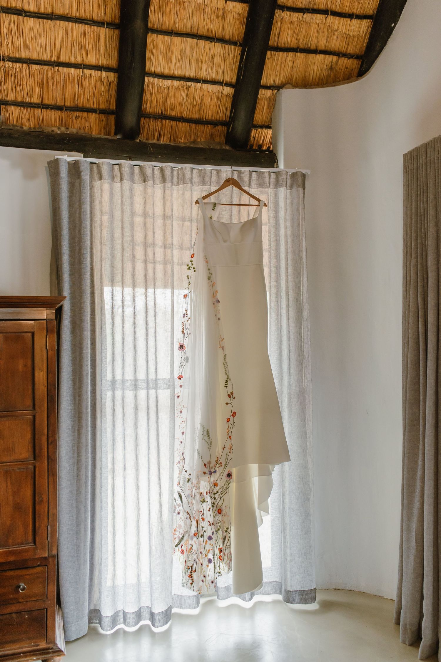 White curtained windows under a thatched roof with wooden beams and rustic furniture.