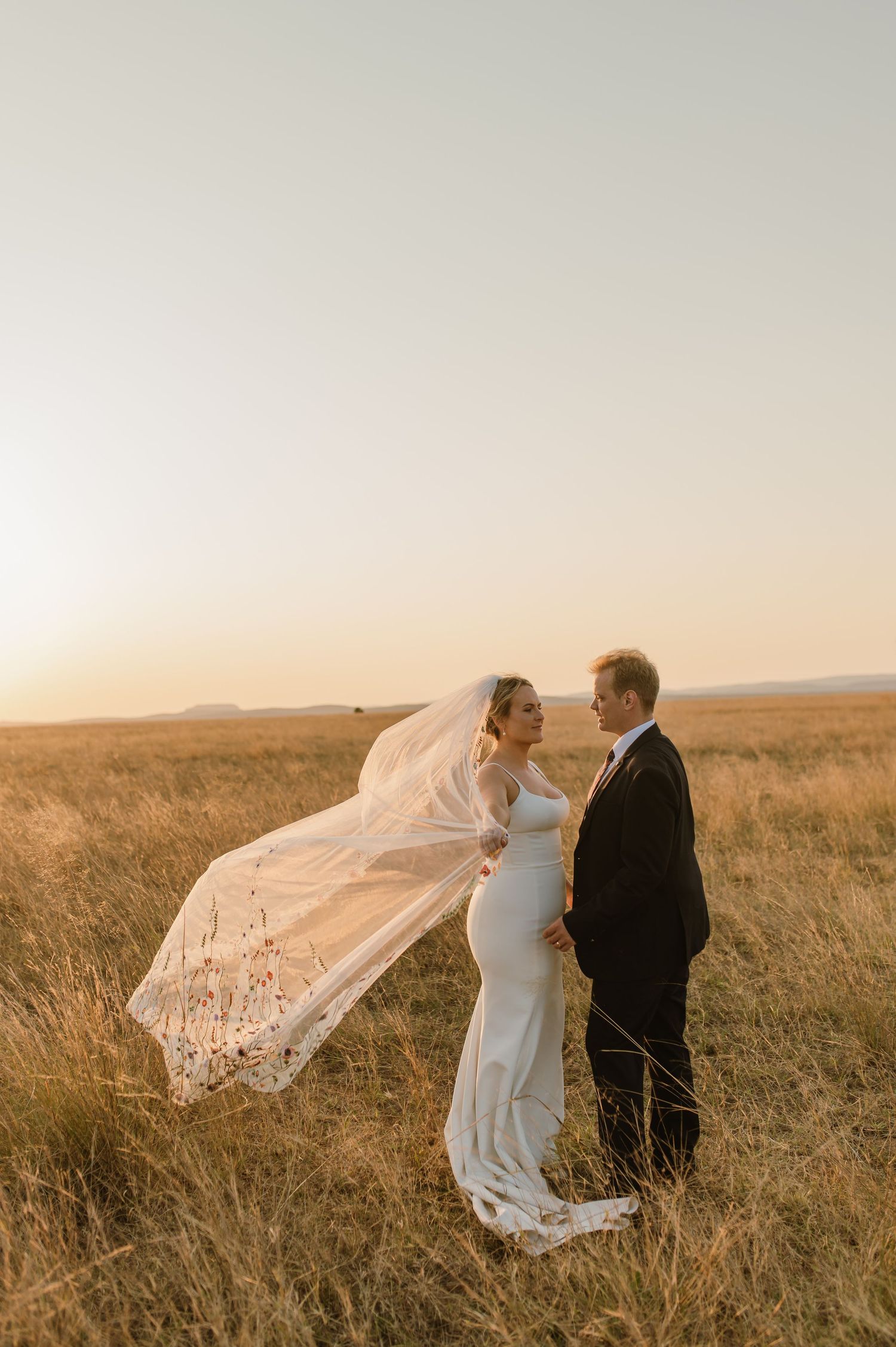 Bride's veil flows dramatically in wind as couple poses in sunset meadow.