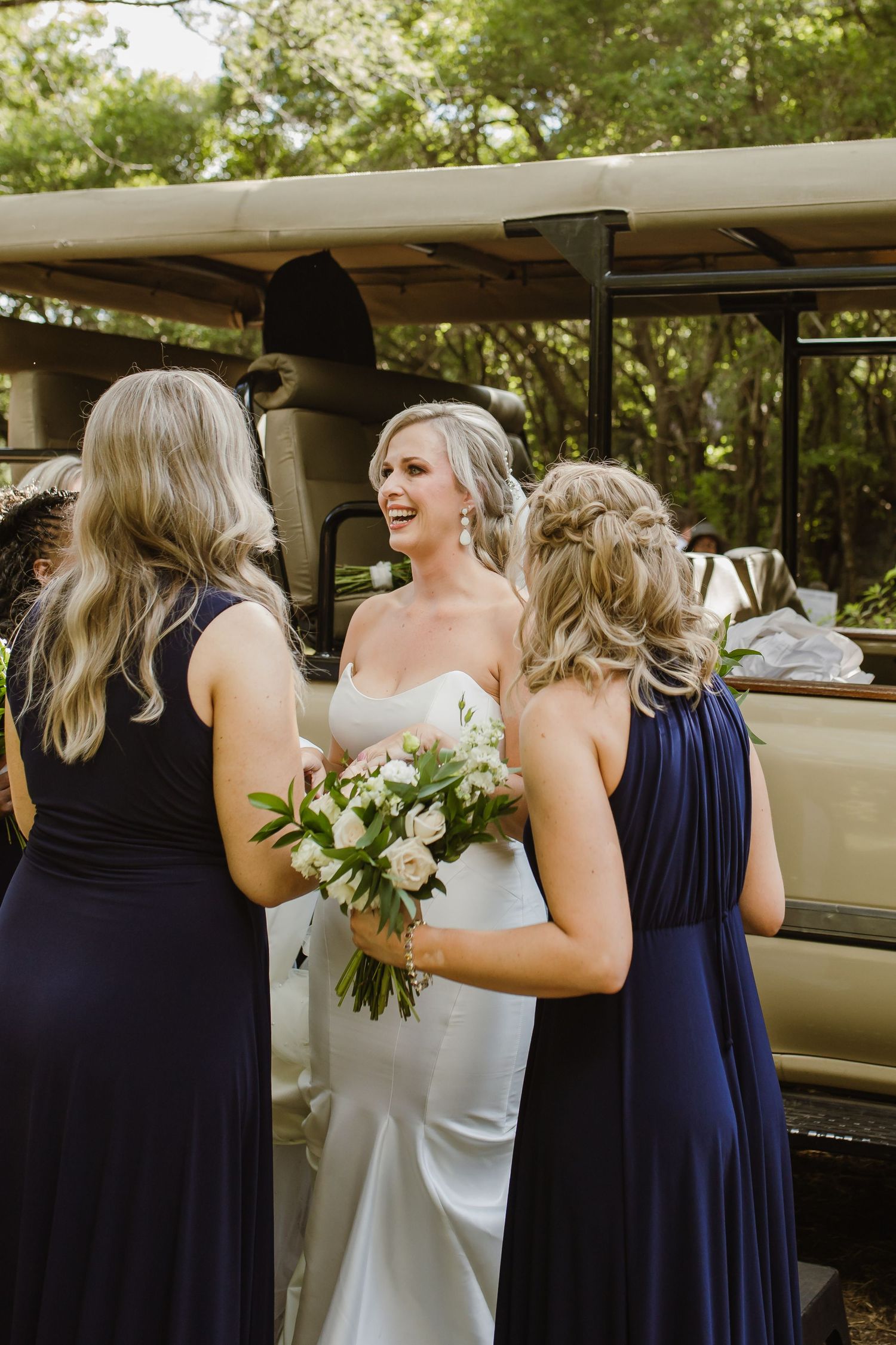 Bridesmaids in navy dresses gather around a white-clad figure near a vintage car during an outdoor wedding.