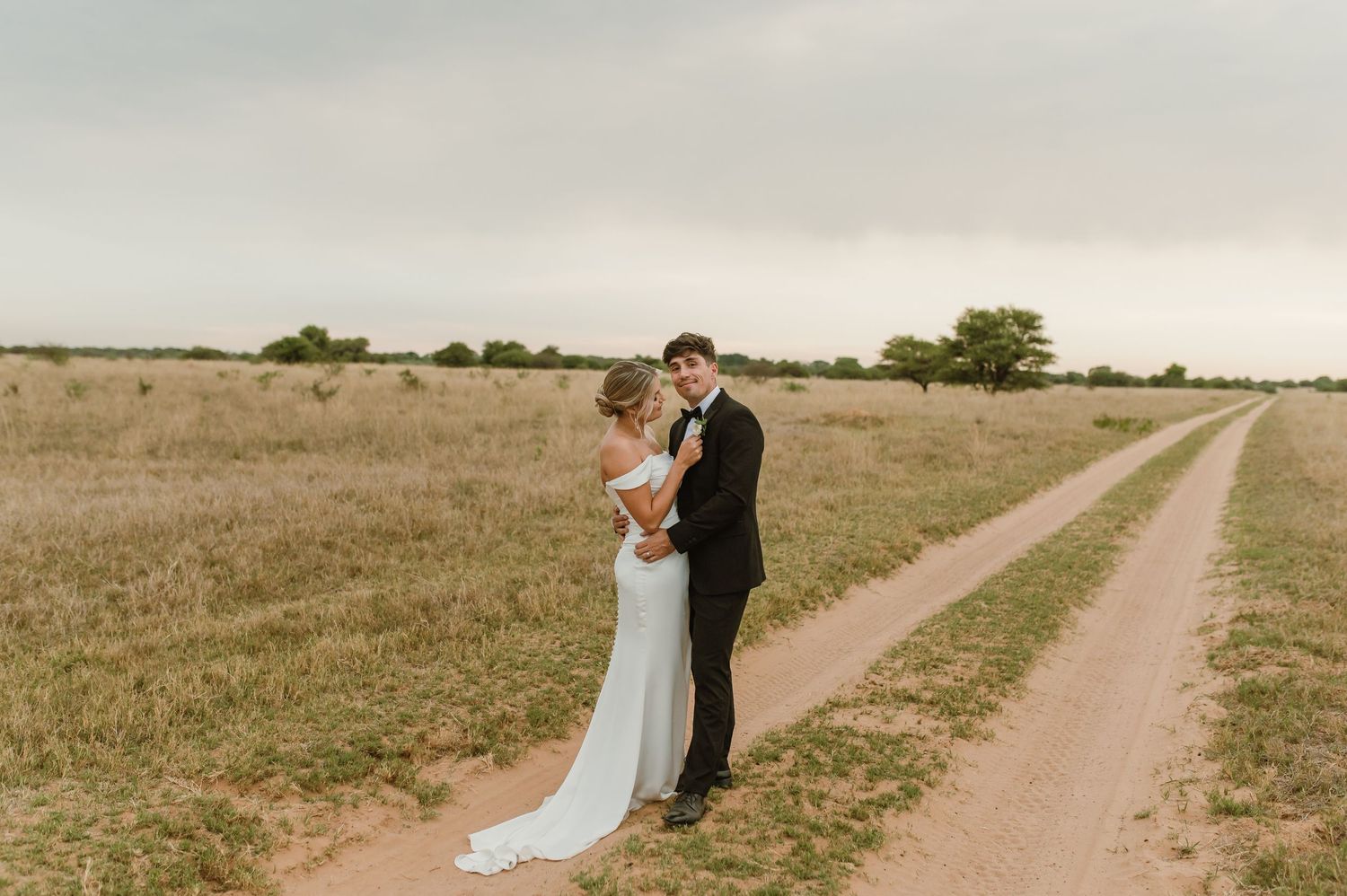 A romantic couple embraces on a dirt path through golden grasslands at sunset.