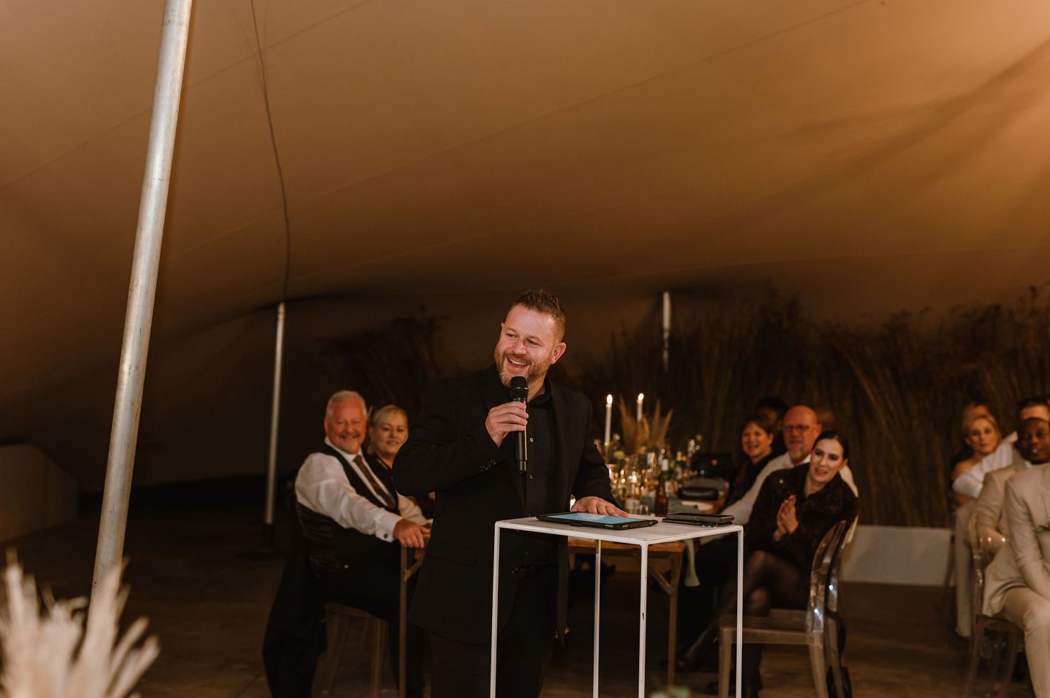 Wedding reception speeches being given under warm lighting in an elegant tent setting.