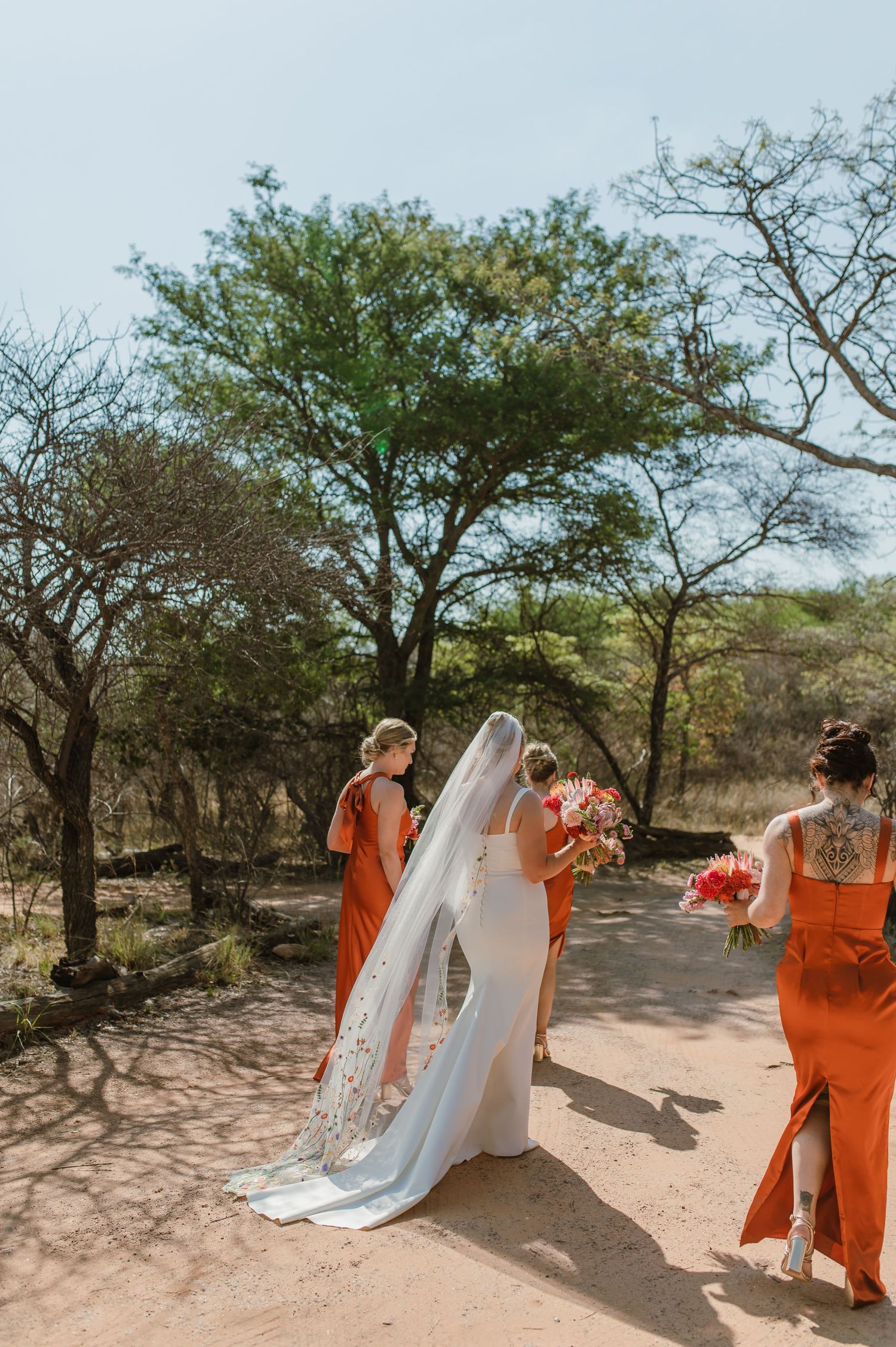 Wedding party in orange dresses walking along desert path with bride in white.