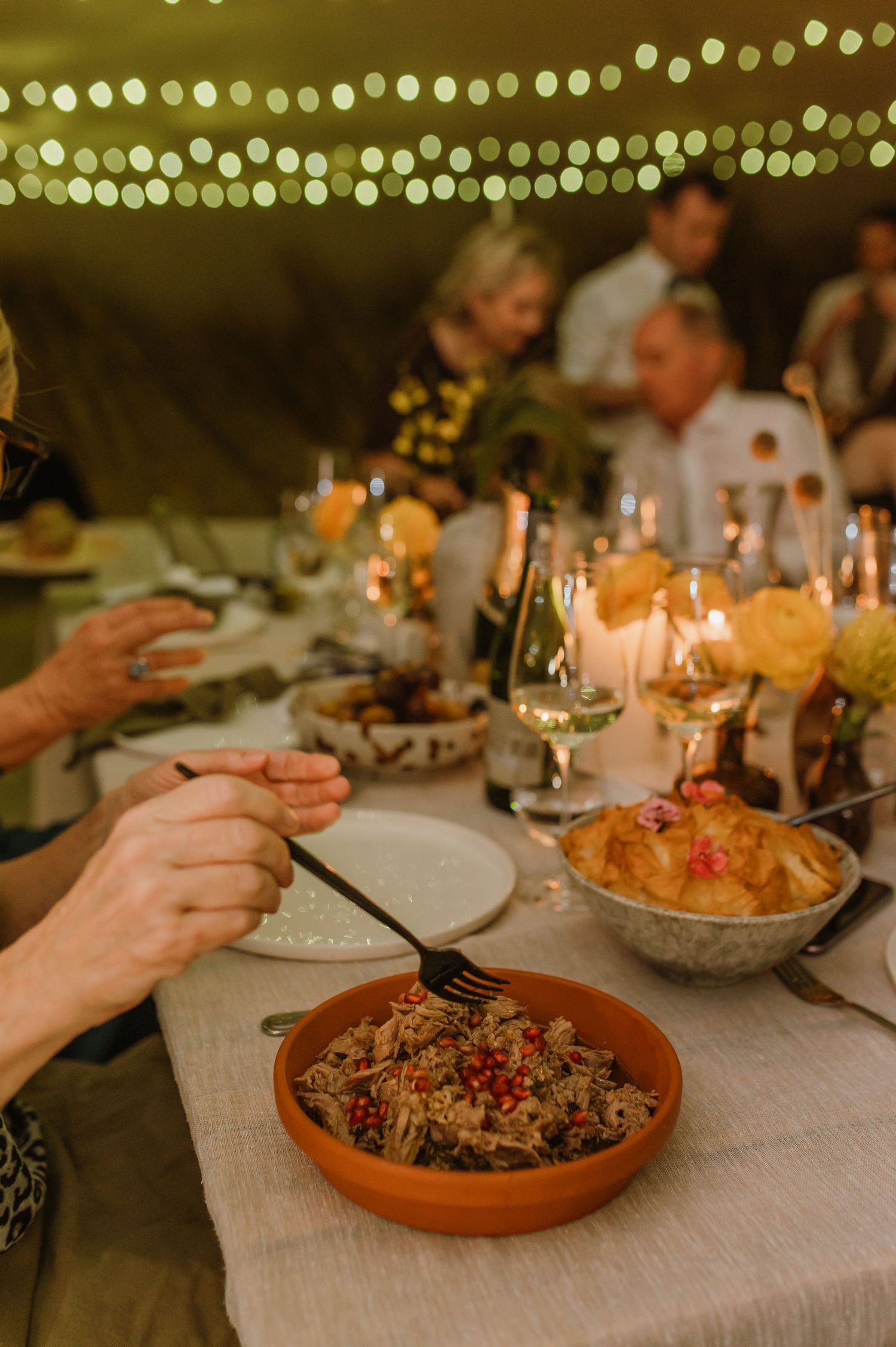 Wedding reception dinner plates with colorful dishes and ambient string lighting overhead.