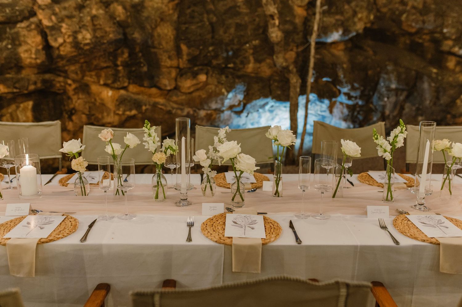 Long banquet tables with white linens and place settings arranged outdoors under natural rock formations.