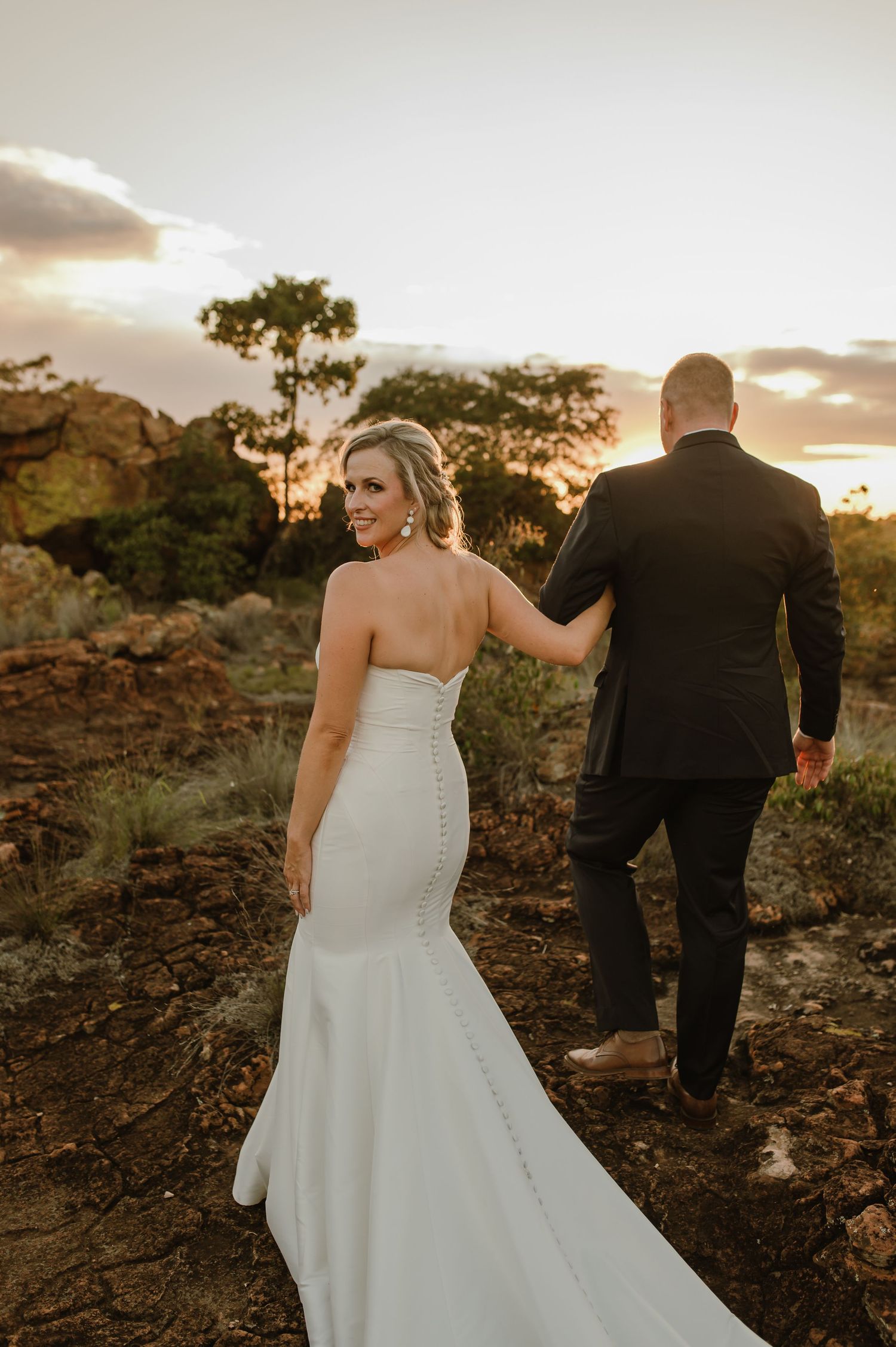 Bride's dress trails behind as couple walks through rocky terrain at sunset.