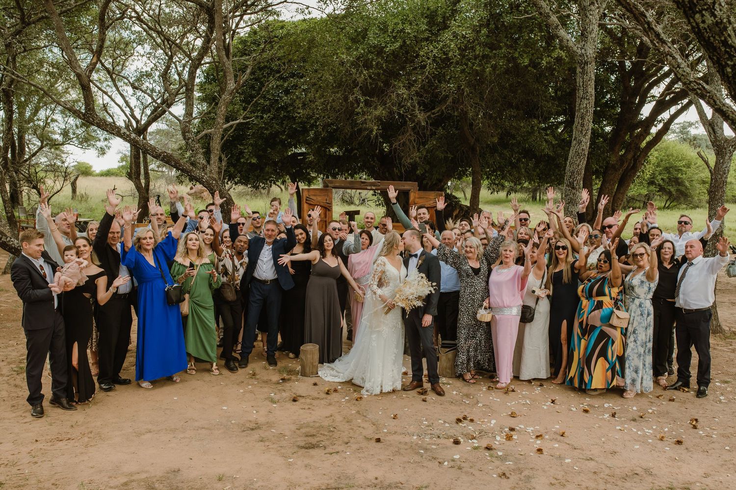 Large wedding group photo with guests gathered outdoors under trees.