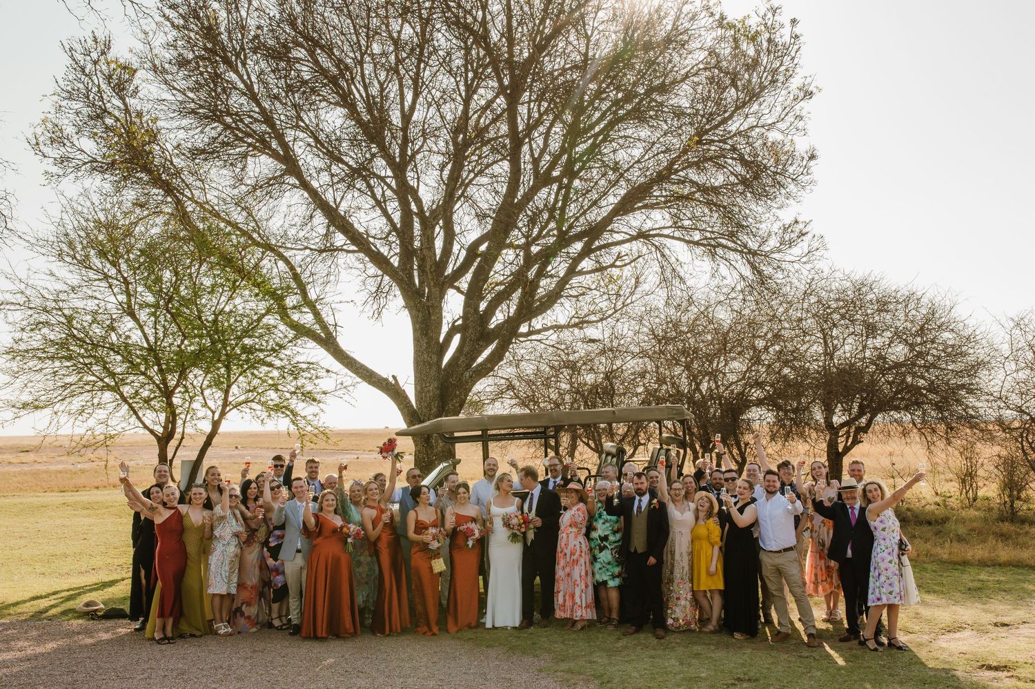 Large wedding party poses for group photo under trees at golden hour.
