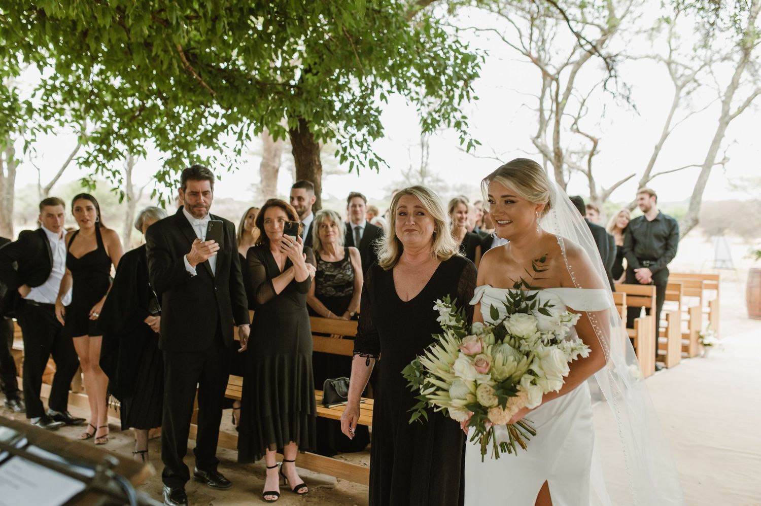 Emotional wedding ceremony moment with guests watching under tree canopy.