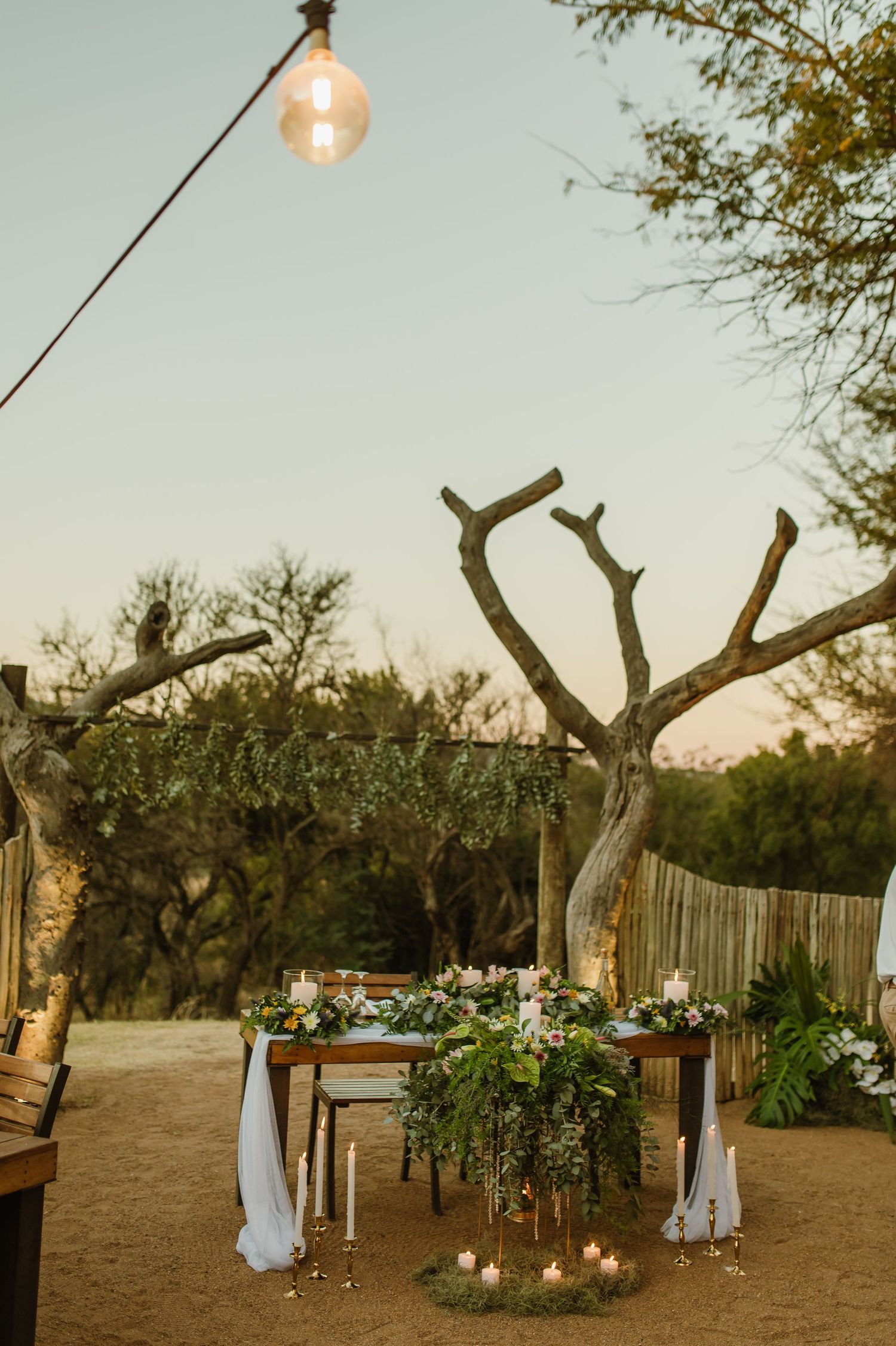 Rustic outdoor wedding venue with string lights, white drapes, and floral decorations at dusk.