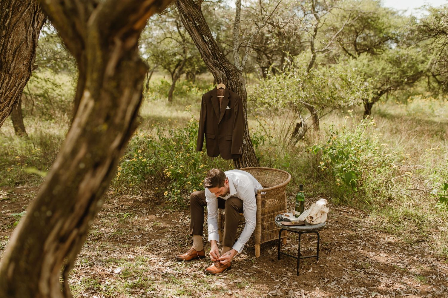 Person getting dressed in an outdoor setting with a brown suit jacket hanging from a tree branch.