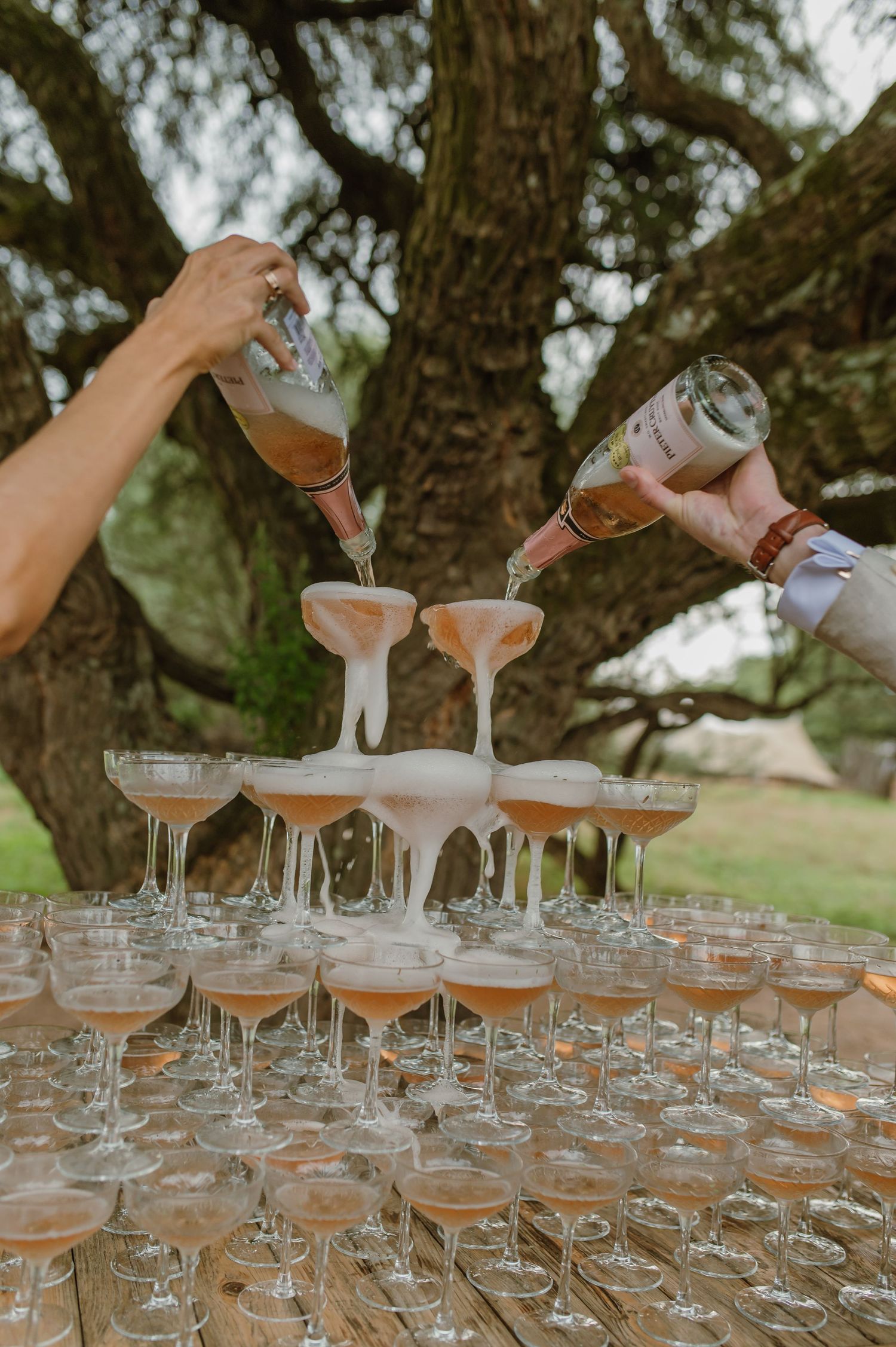 Hands pouring drinks into multiple champagne glasses at an outdoor wedding bar.