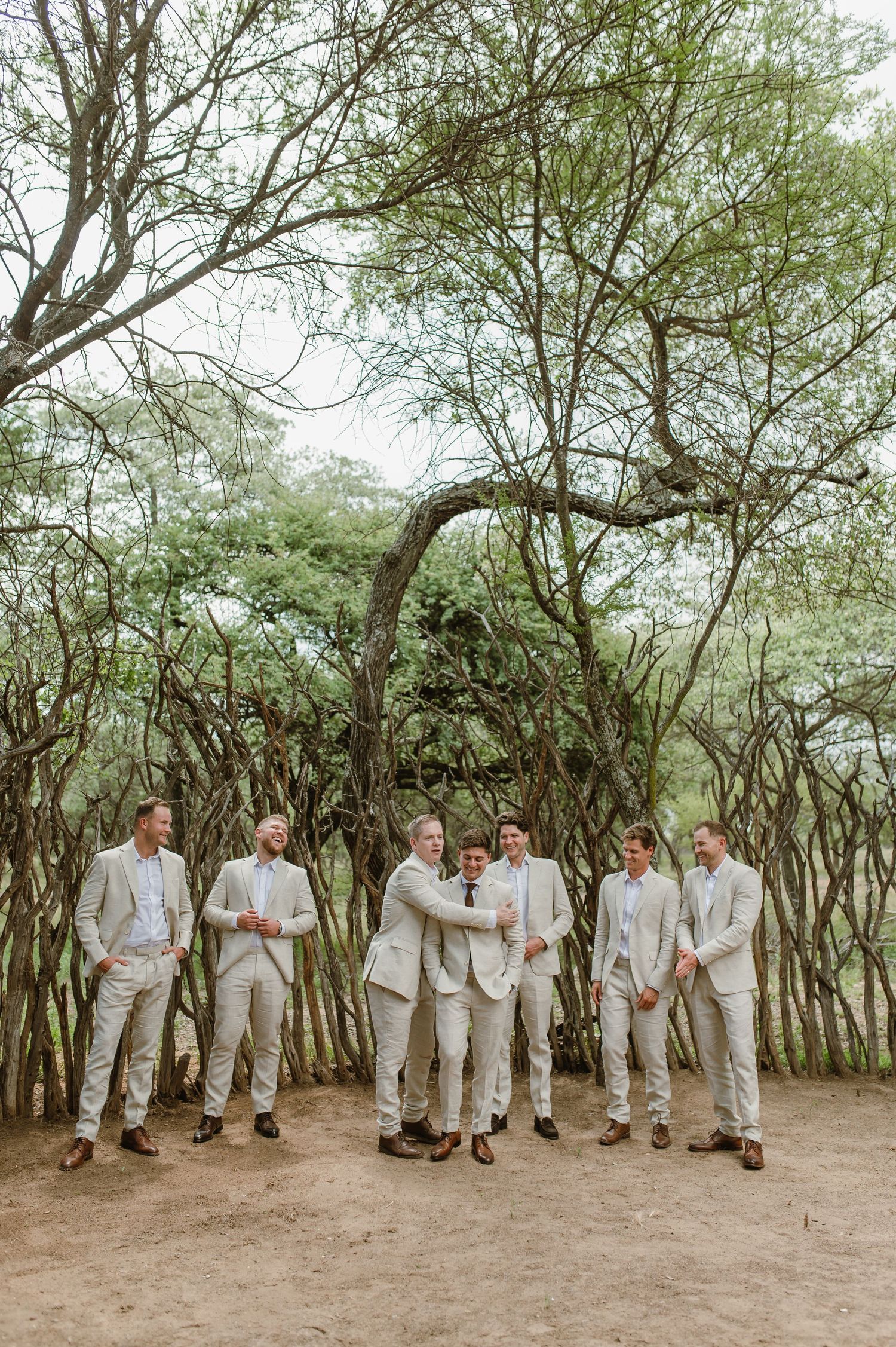 Wedding party members in light colored suits stand together on a dirt path surrounded by trees.