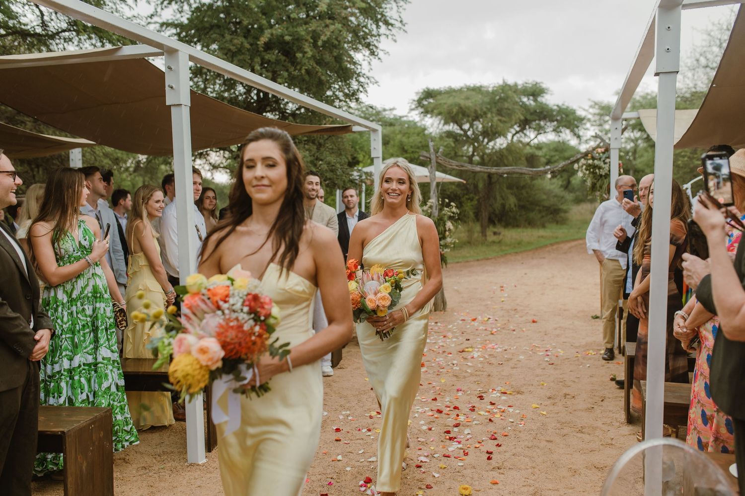 Wedding party members walk down a dirt path lined with scattered flower petals.
