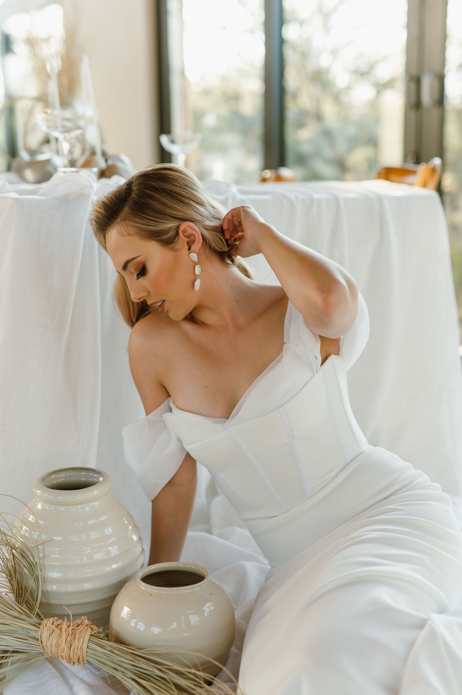 An elegant wedding scene with a white off-shoulder dress and ceramic vases in a light-filled room.