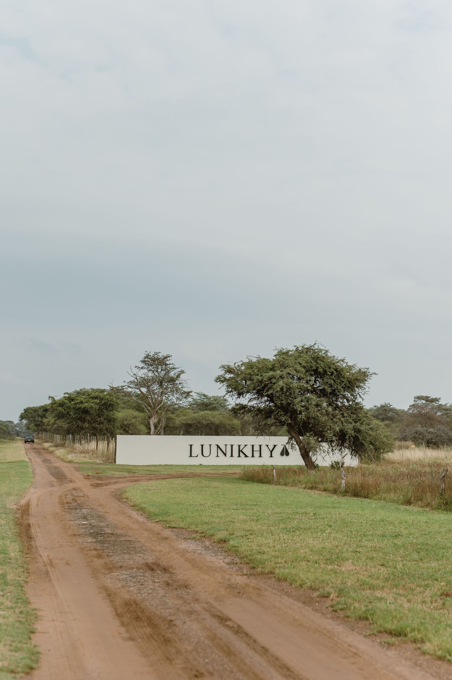 A dirt road stretches through rural African grassland with scattered trees and a Lunikhya sign visible in the landscape.