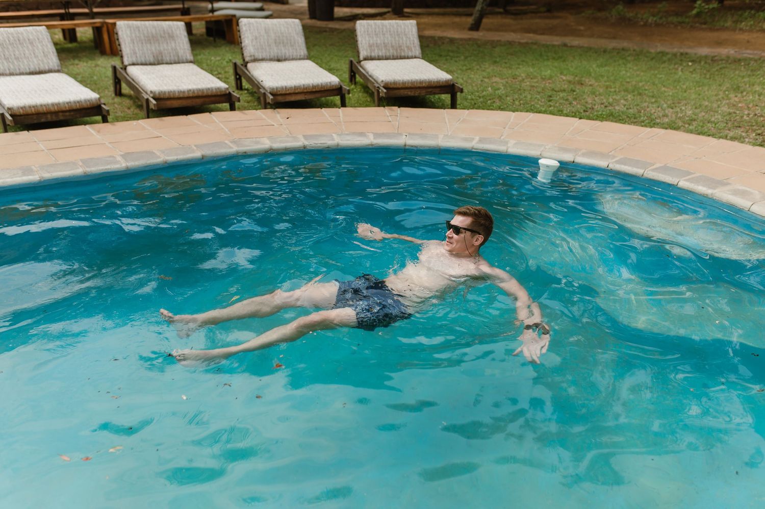 Someone floats peacefully on their back in a turquoise swimming pool with lounge chairs visible in the background.