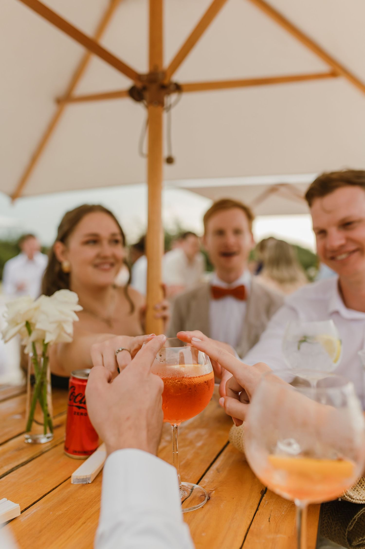 Wedding guests enjoy colorful cocktails at a wooden table under a white umbrella canopy.