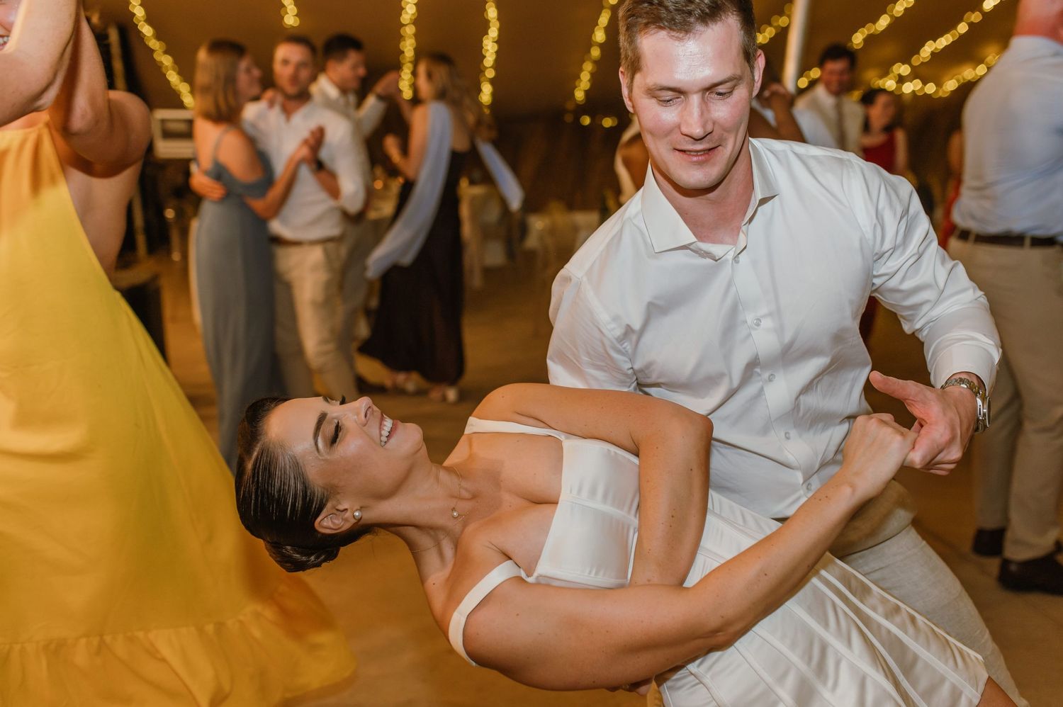 Couple performs dramatic dance move during wedding reception under string lights.