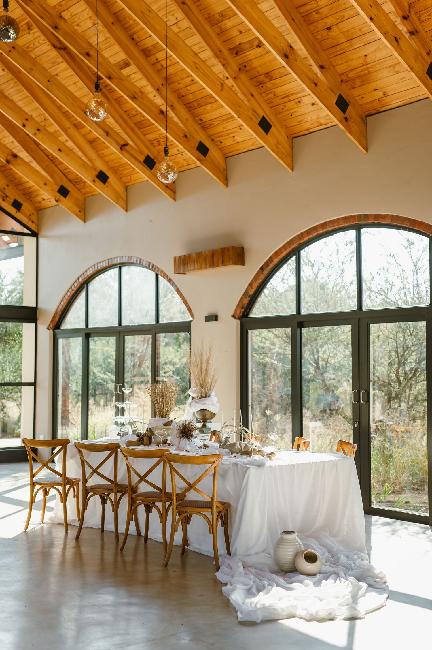 Spacious venue interior with wooden ceiling beams, arched windows, and white table settings.