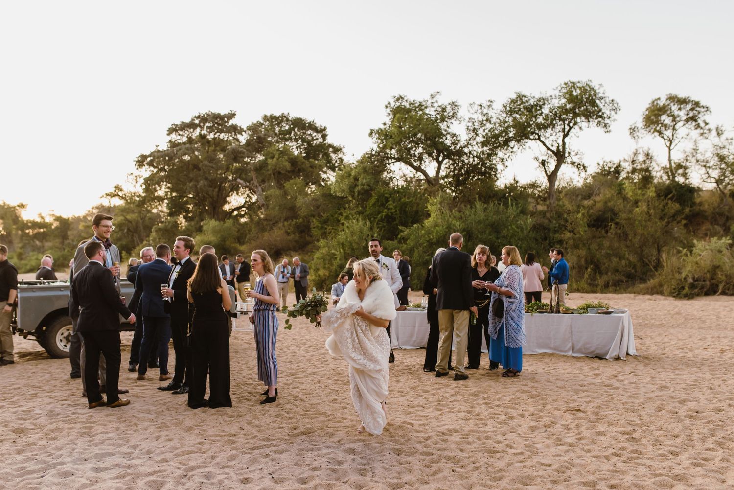 Wedding guests gather on a sandy beach path at sunset for an outdoor ceremony.