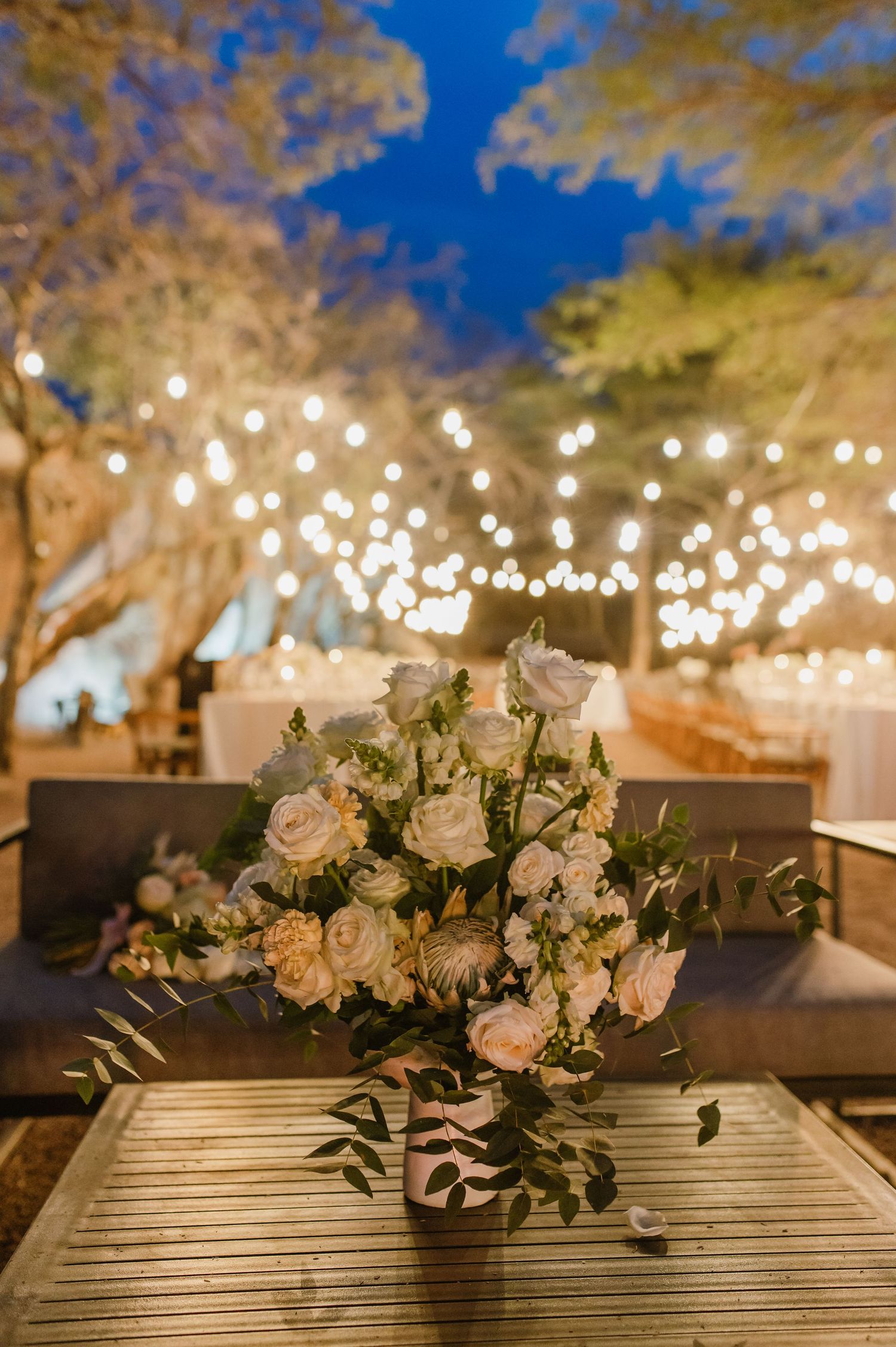 White floral centerpieces on wooden tables under string lights at outdoor wedding reception.