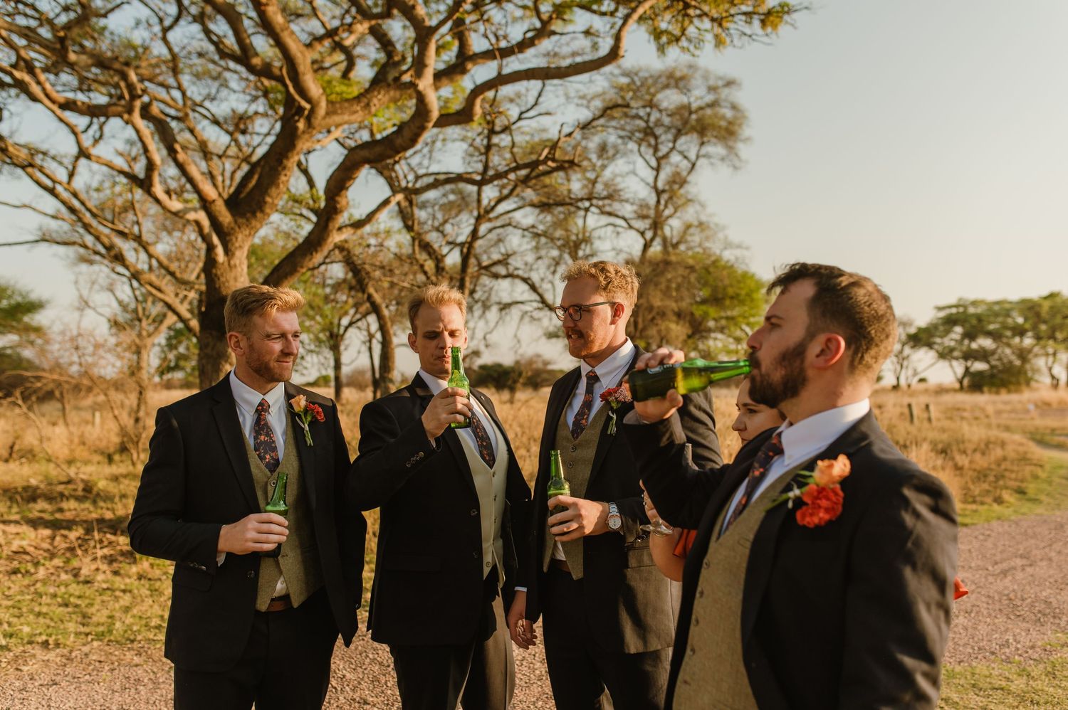 Groomsmen in dark suits toast and celebrate outdoors under autumn trees.