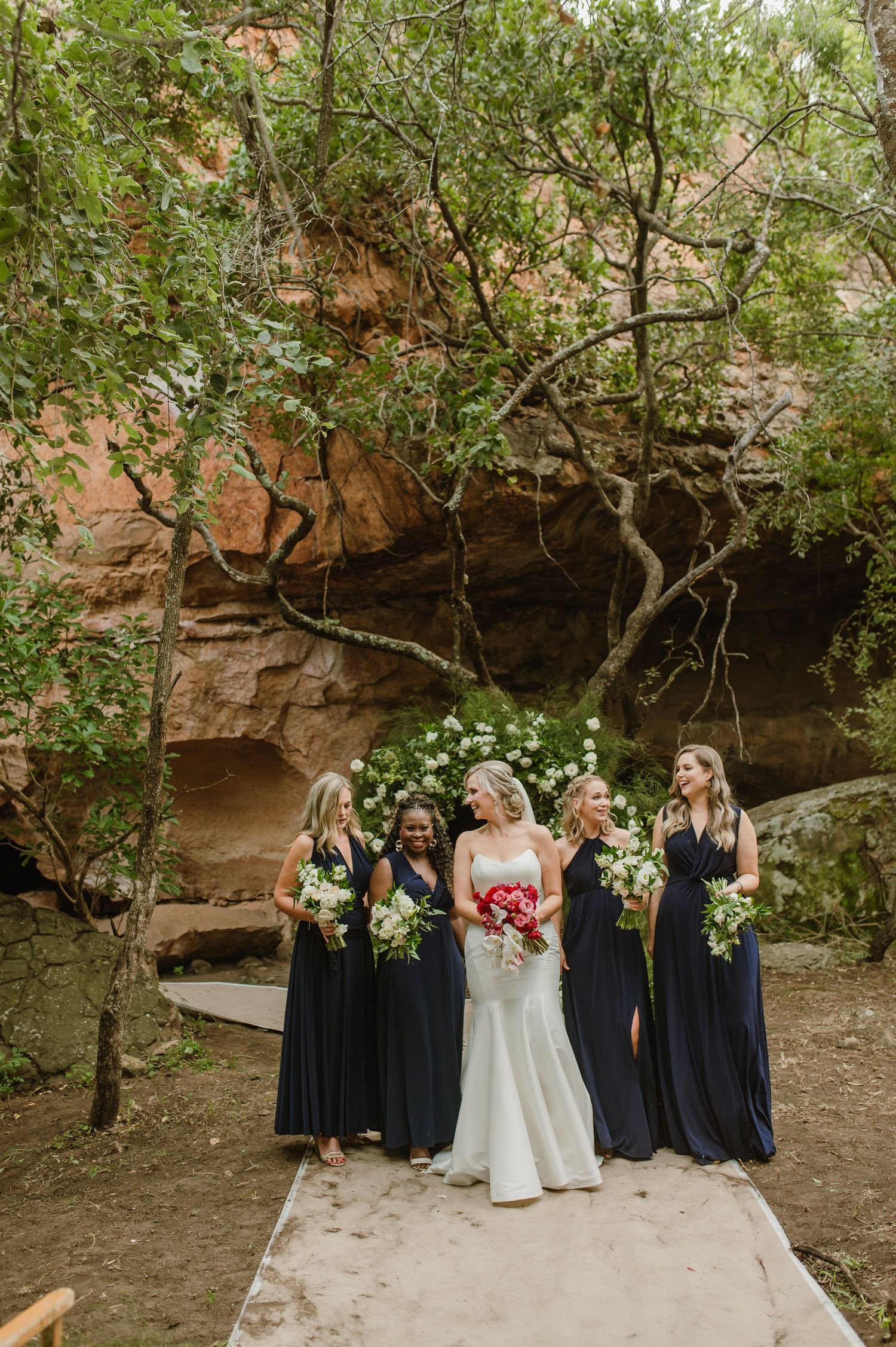A bridal party poses together against red rock formations wearing navy blue dresses and holding bouquets.
