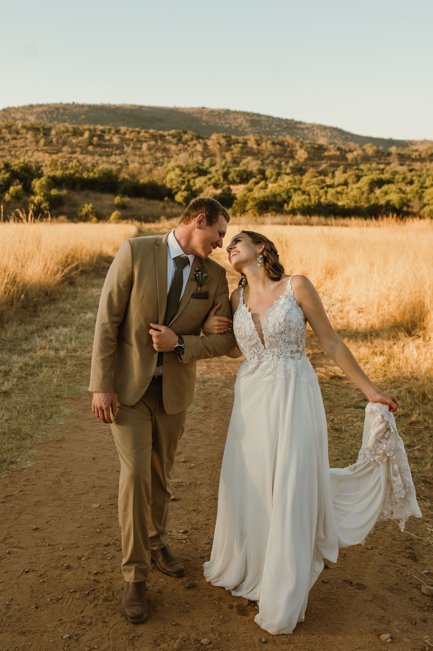Couple shares intimate moment while walking on dirt path through golden field at sunset.