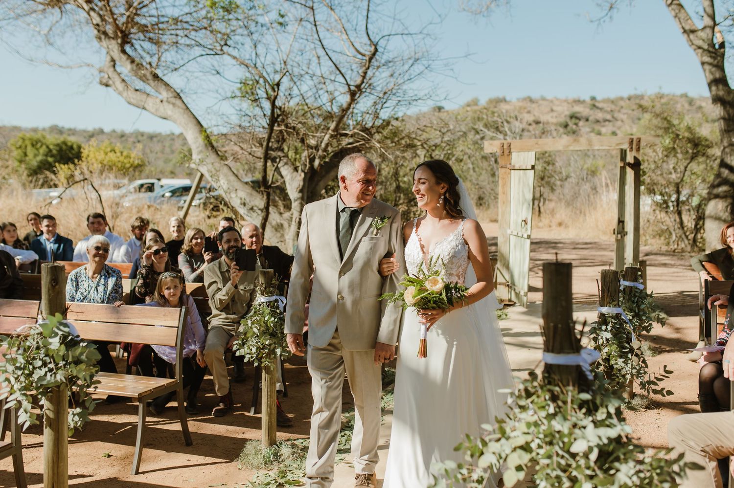 Outdoor desert wedding ceremony with guests gathered under natural tree canopy.