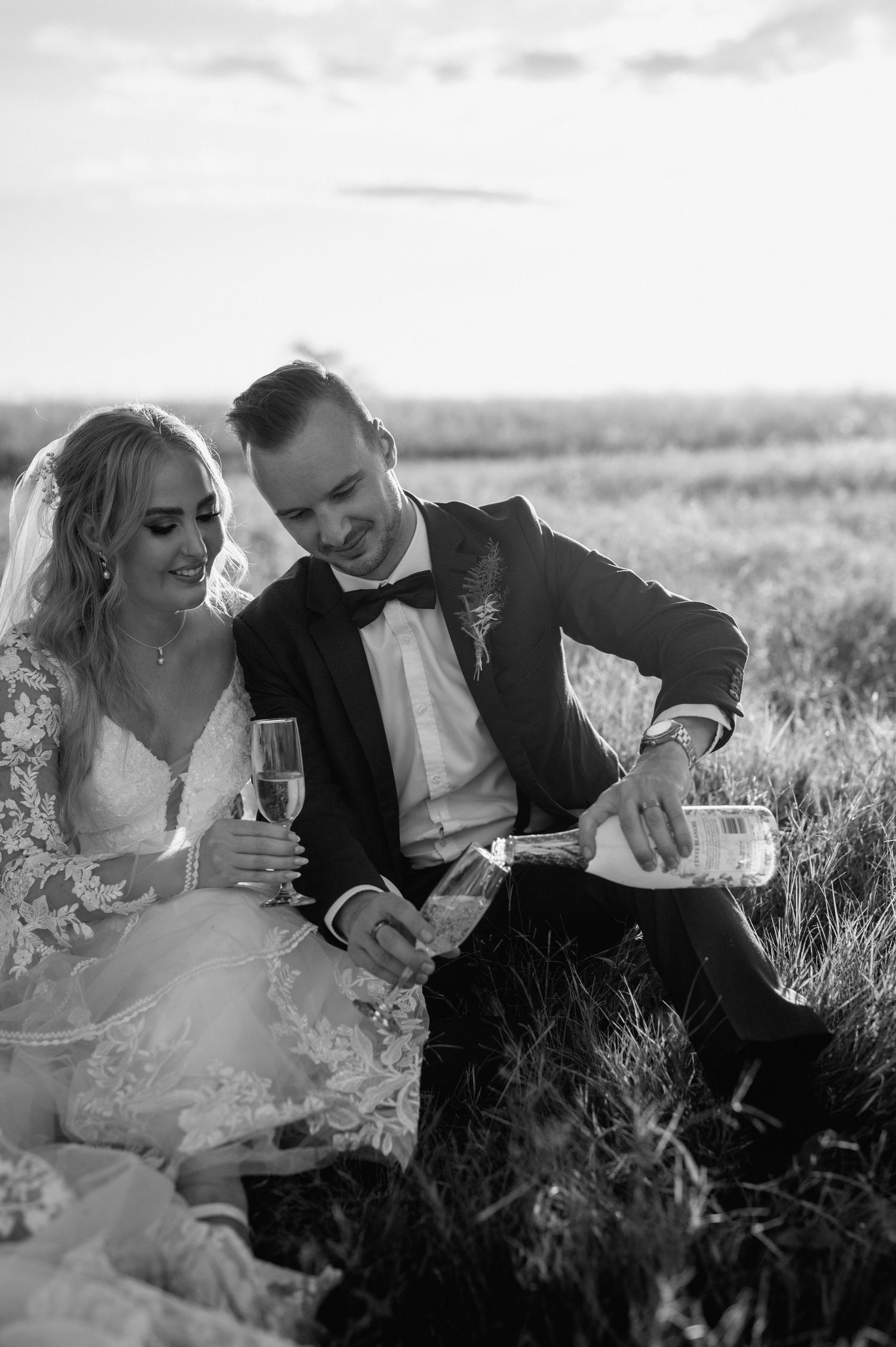 A black and white photo captures a couple toasting with champagne in formal wedding attire.