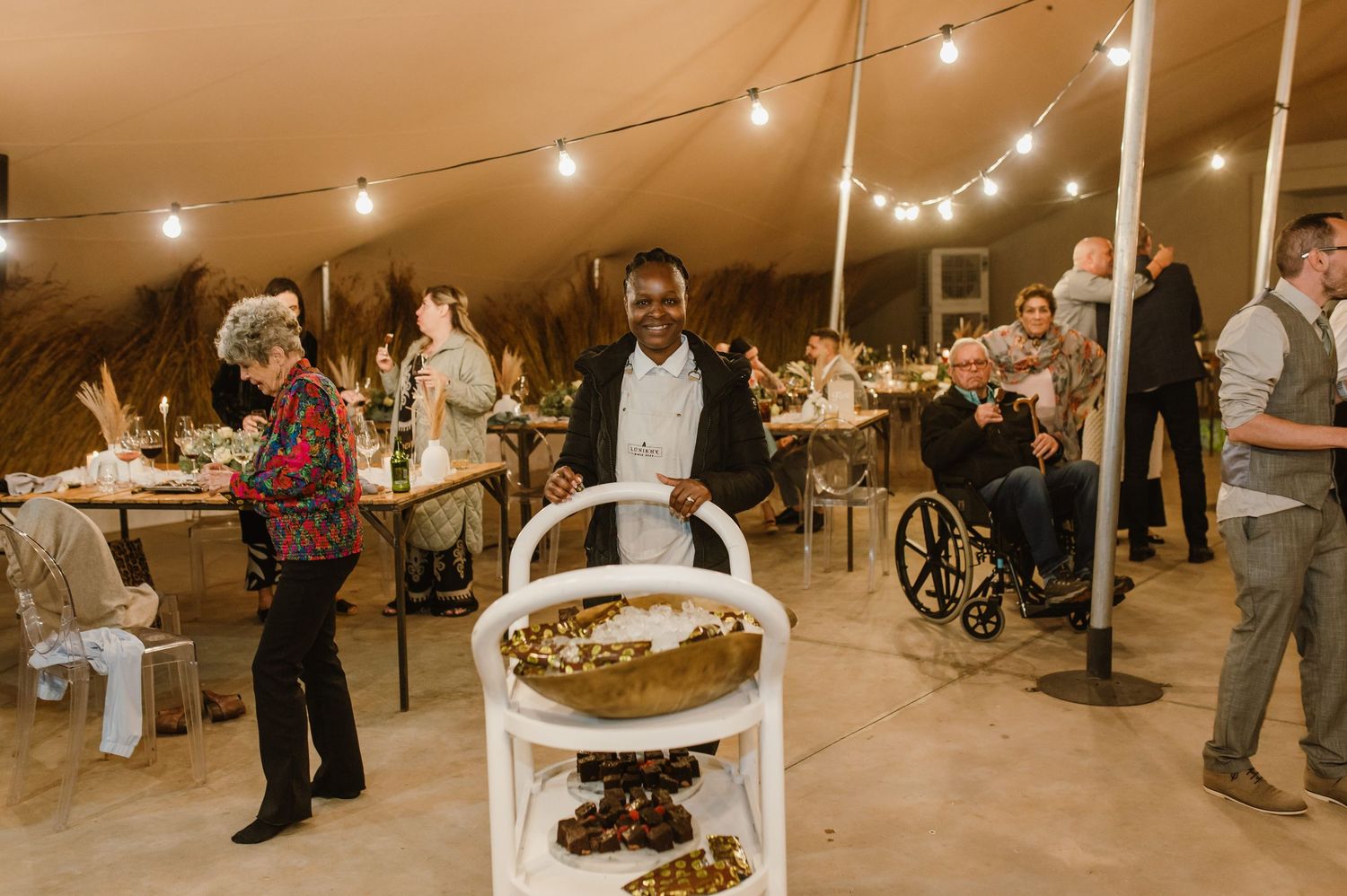 Wedding reception under a tent with string lights and guests mingling around dessert cart.